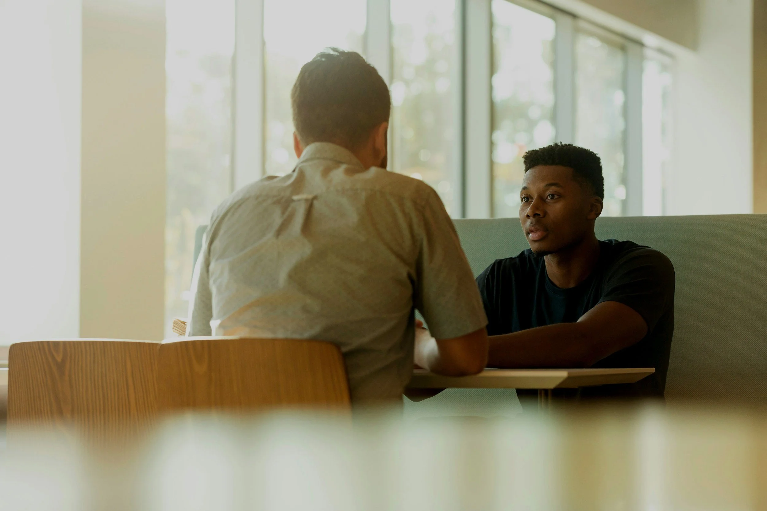 Two men are sitting across from each other in a brightly lit room with large windows, engaged in conversation.