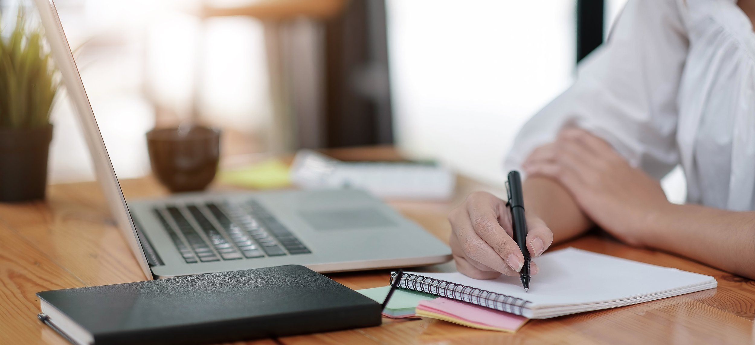 Person writing in a notebook at a wooden desk with a laptop, closed black notebook, sticky notes, and potted plants, in a bright room.