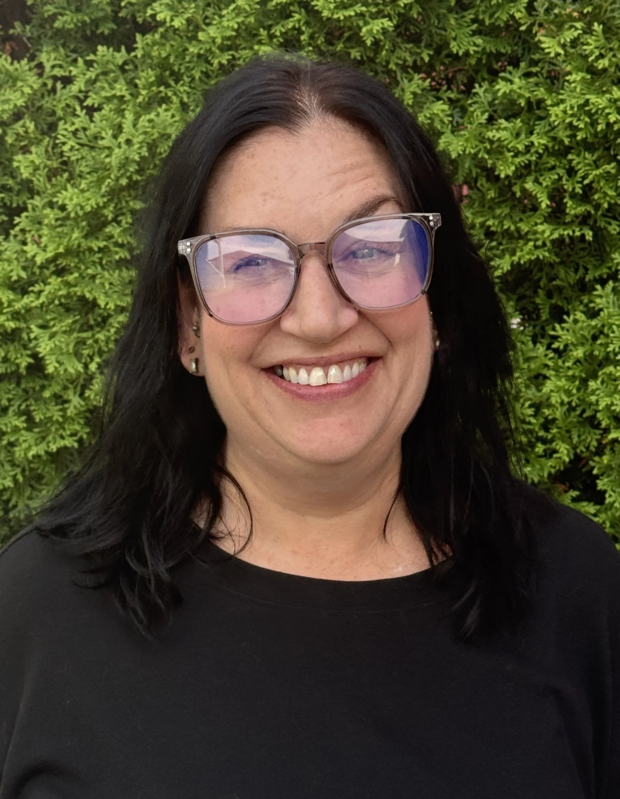 A woman with dark hair, wearing sunglasses and earrings, smiling, standing in front of green foliage.