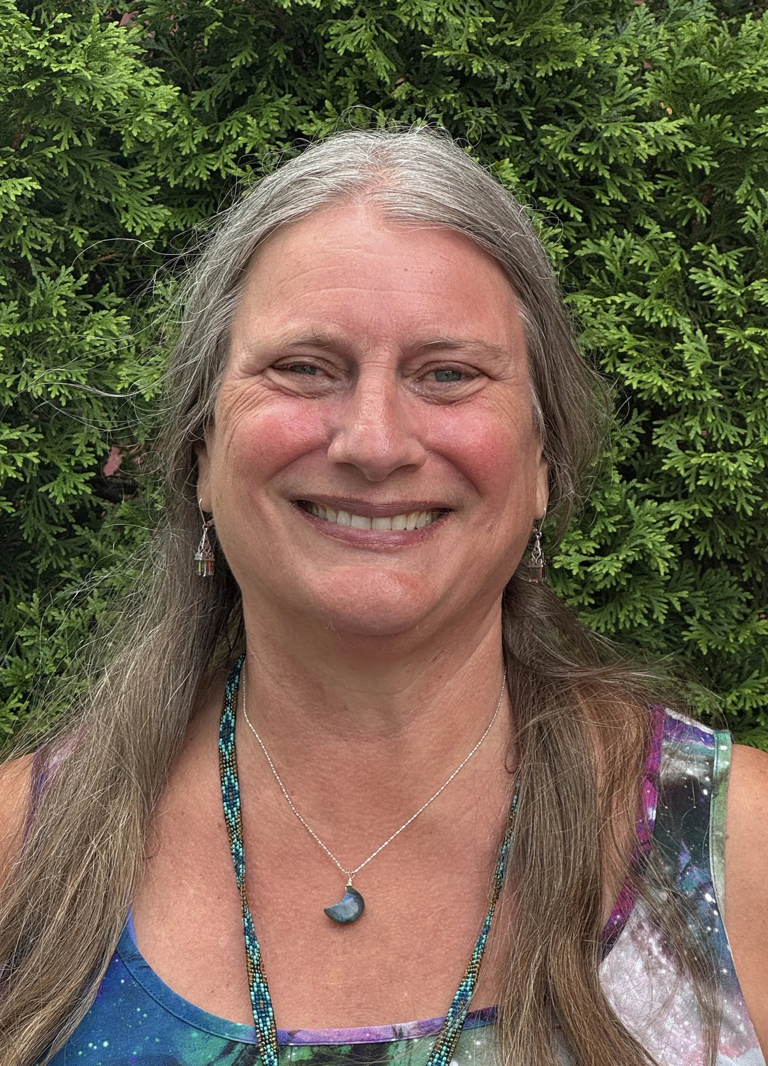 Close-up photo of a smiling woman with long gray hair, wearing colorful jewelry and a galaxy-themed top, standing in front of a green leafy bush.