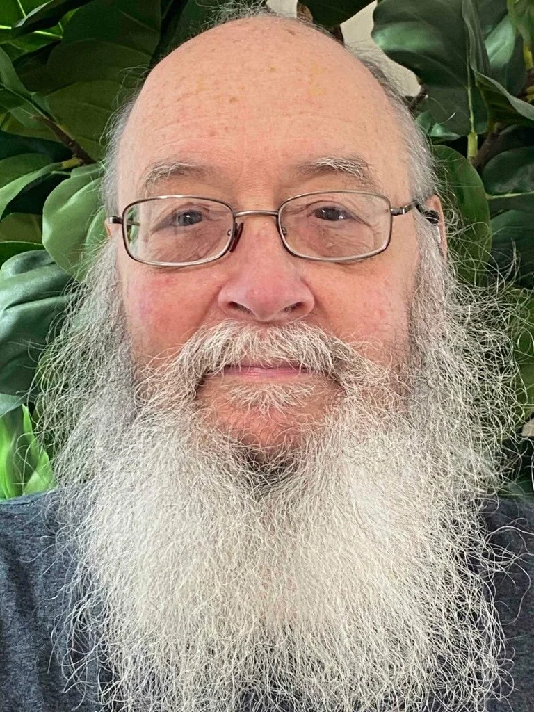 Close-up of an elderly man with glasses and a long white beard, standing in front of green leafy plants.