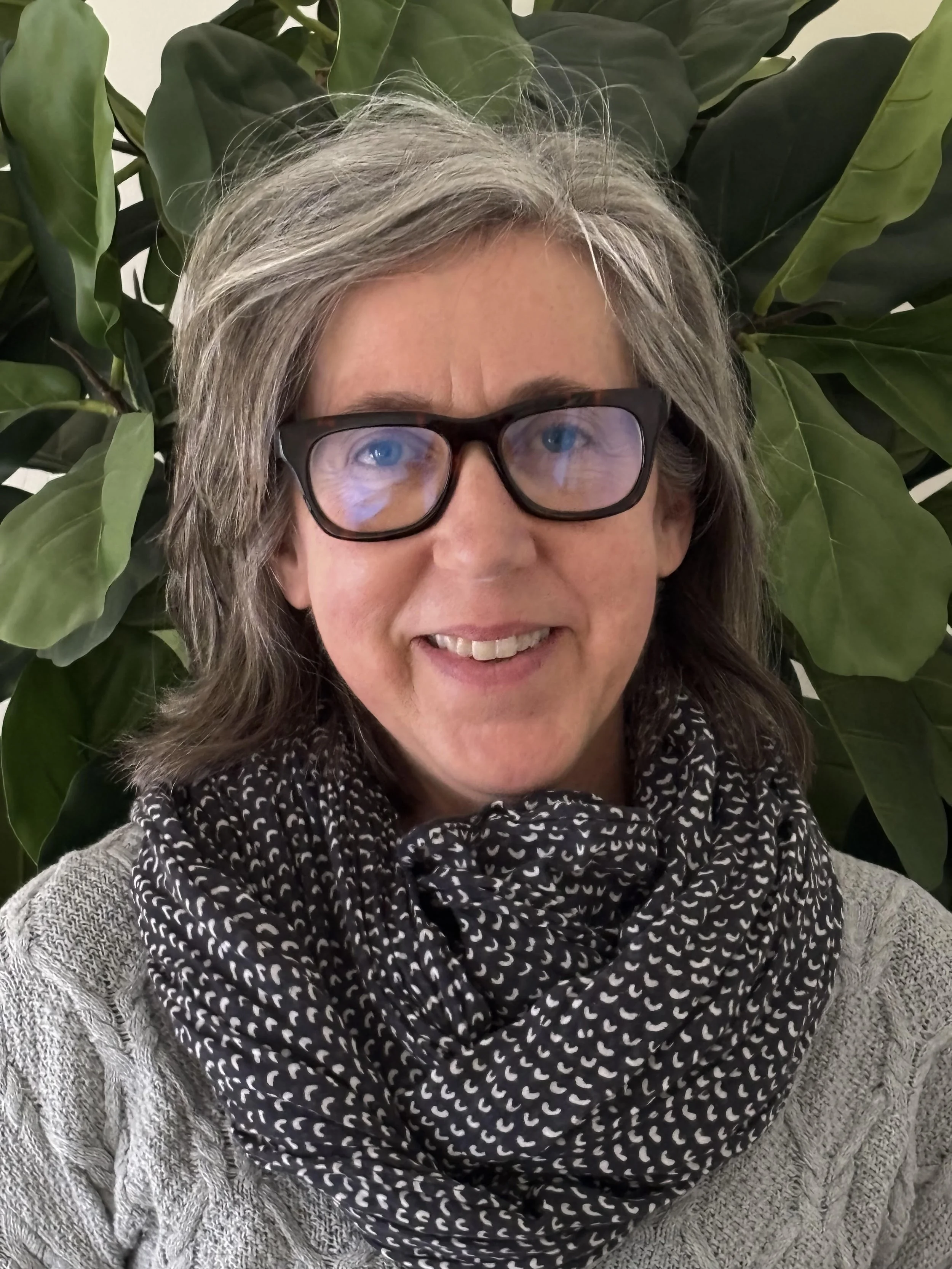 A woman with glasses and gray hair smiling, sitting in front of large green leaves, wearing a gray sweater and a black and white patterned scarf.