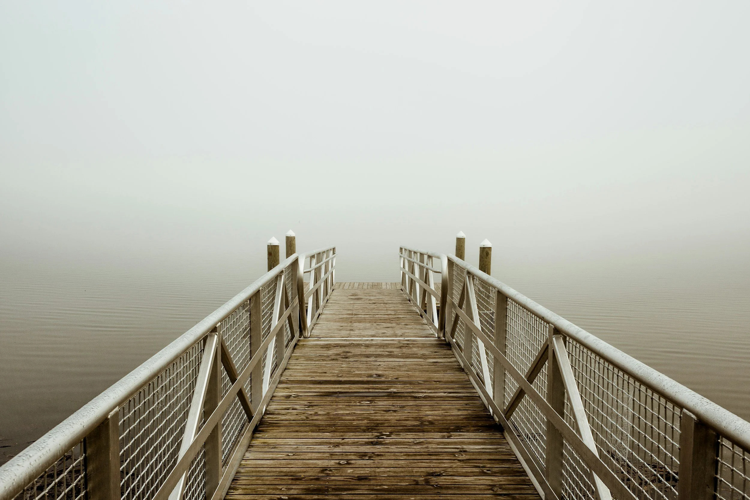 A wooden dock extending over a body of water with foggy at Cayuga Lake with overcast sky and minimal visible background.