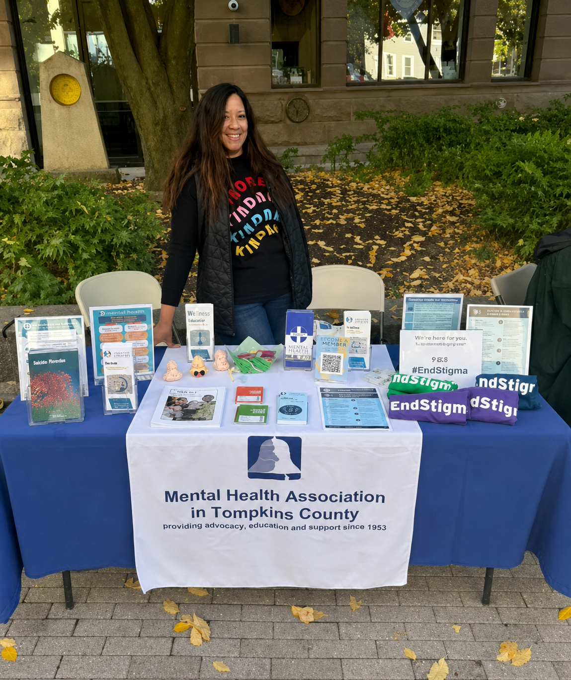 CEO of MHA, Josephine Gibson, stands at an outdoor table organized by the Mental Health Association in Tompkins County, with informational pamphlets and advocacy items related to mental health awareness and support.