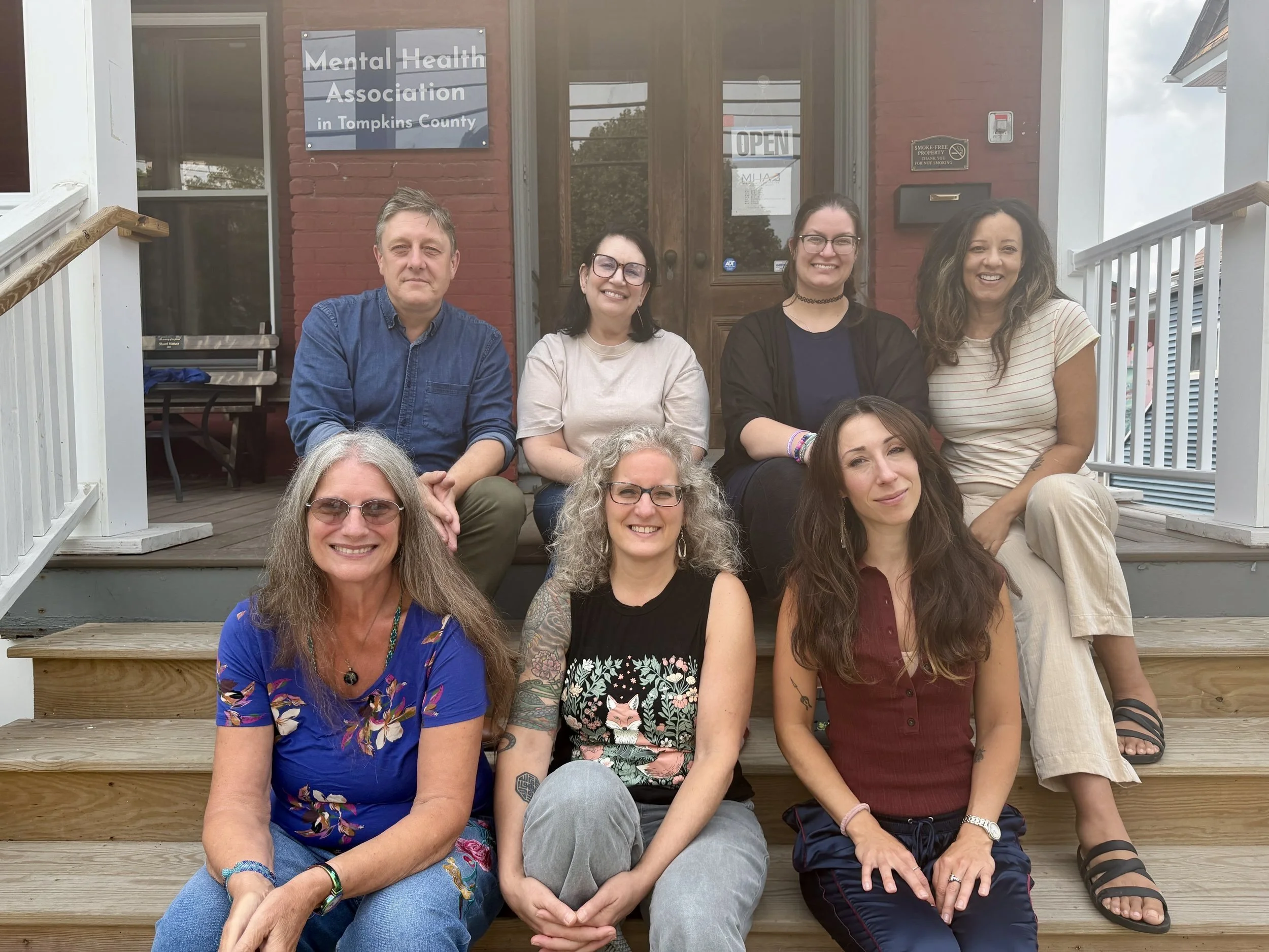 Seven MHA staff sitting on wooden stairs outside, smiling in front of a brick building with a sign reading 'Mental Health Association in Tompkins County,' and an 'OPEN' sign on the door.