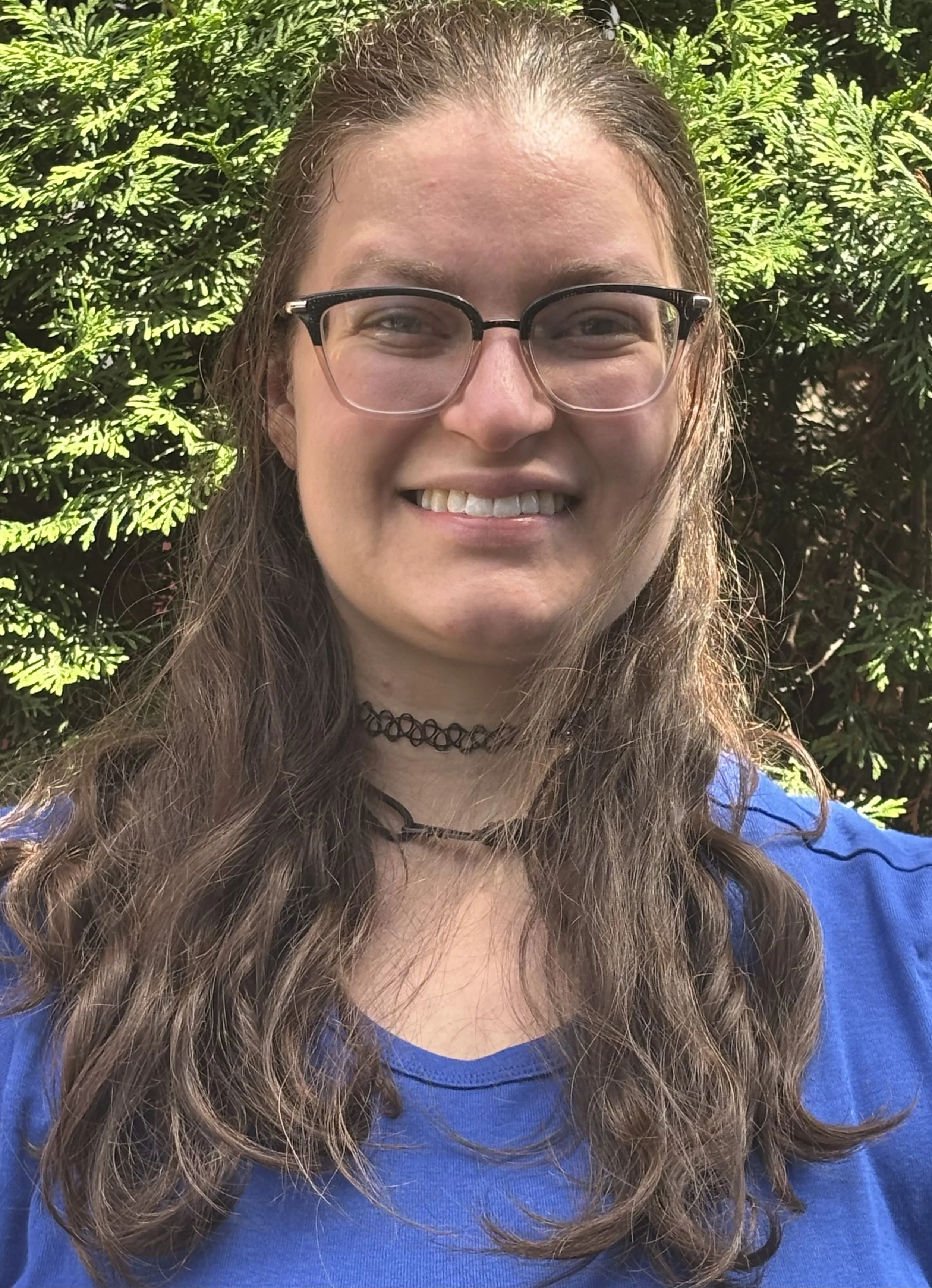 A woman with long, curly brown hair, wearing glasses, a black choker, and a blue shirt, smiling outdoors against green leafy background.