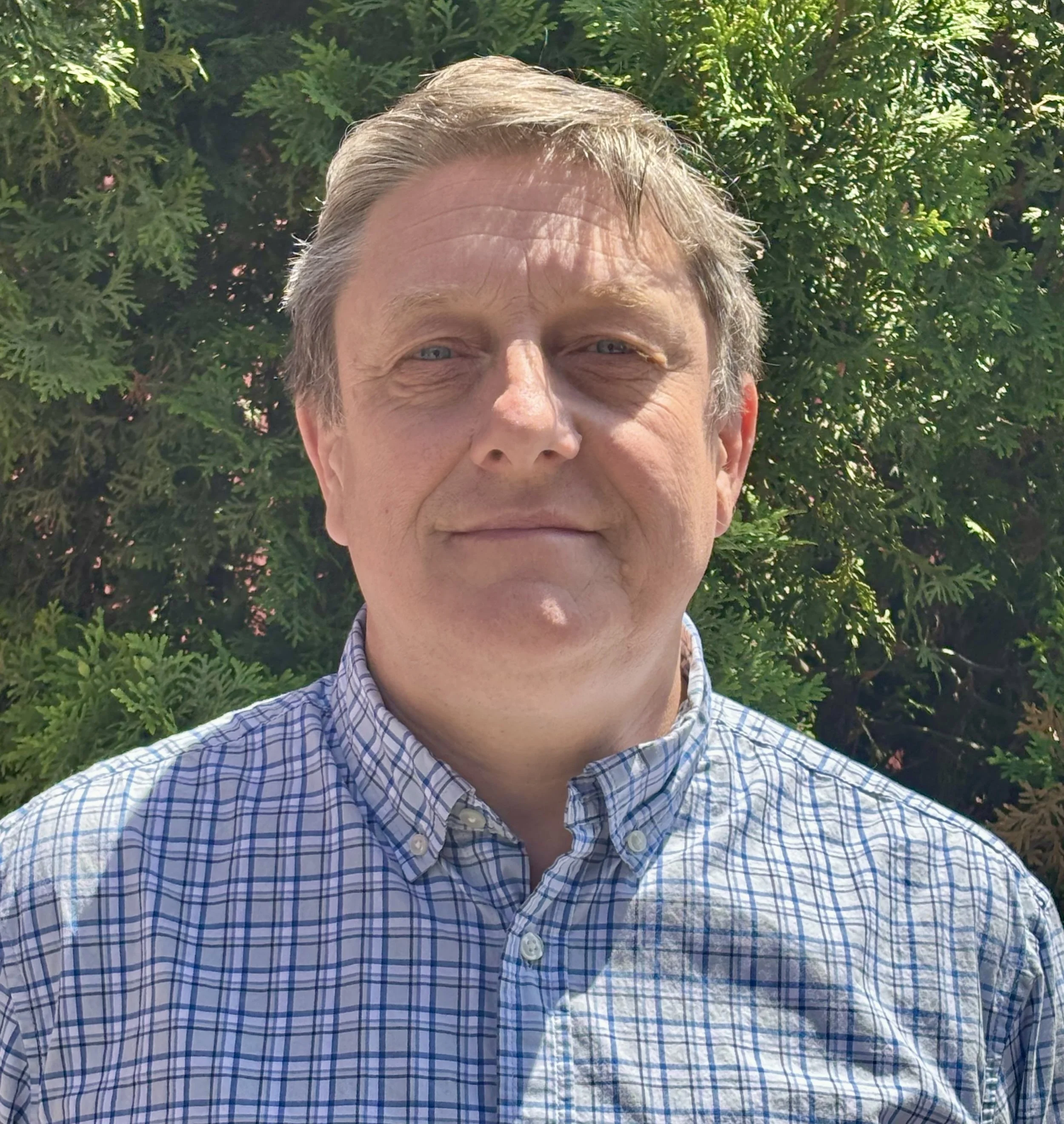 Close-up of a middle-aged man's face with gray hair and blue eyes, wearing a blue and white checkered shirt, standing outdoors in front of green leafy bushes.
