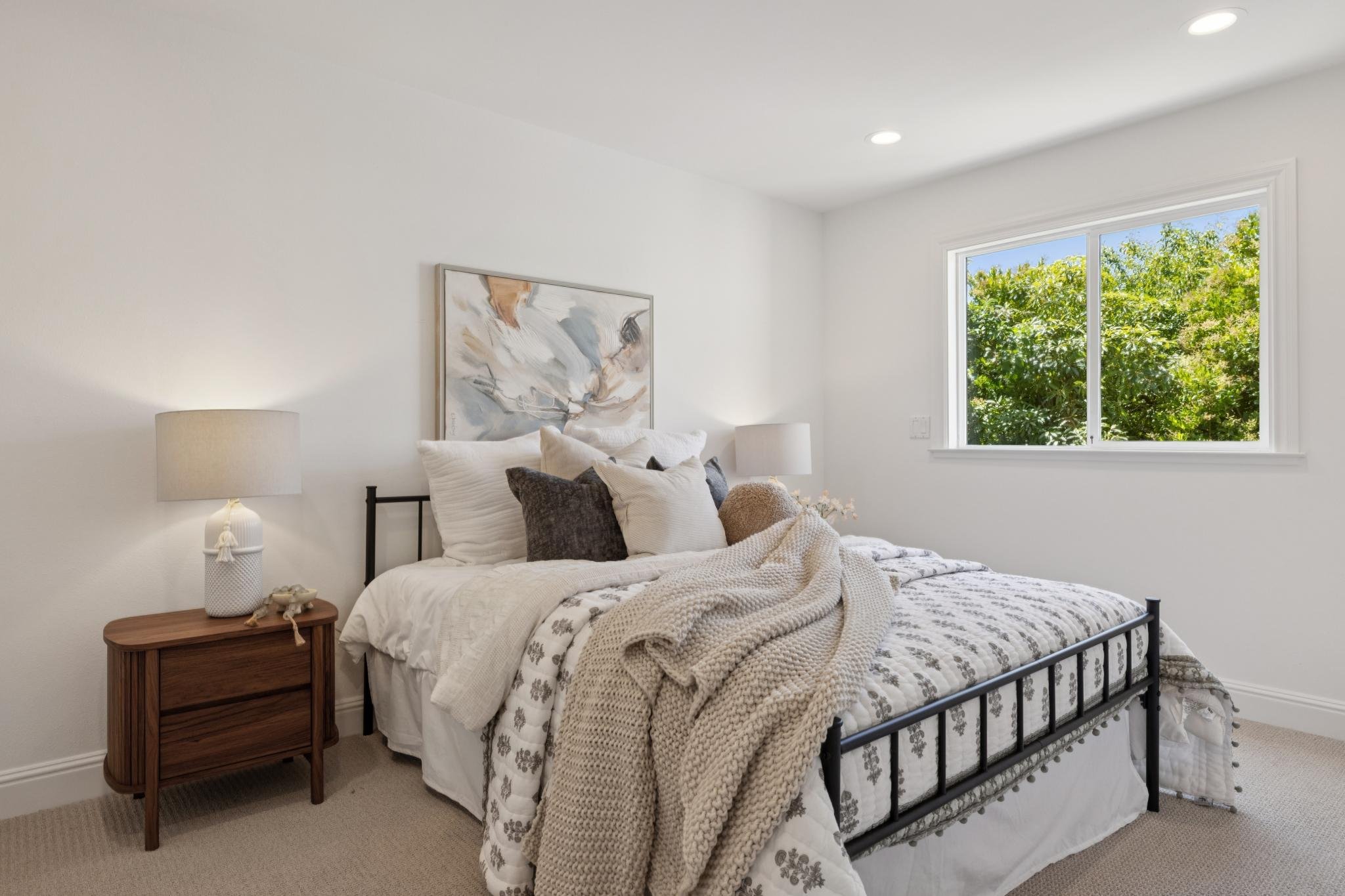 A bedroom with a black metal bed frame, white bedding, and various pillows. There is a wooden nightstand with a white table lamp and small decorative items. A large window shows green trees outside, and a piece of abstract art hangs above the bed. Th