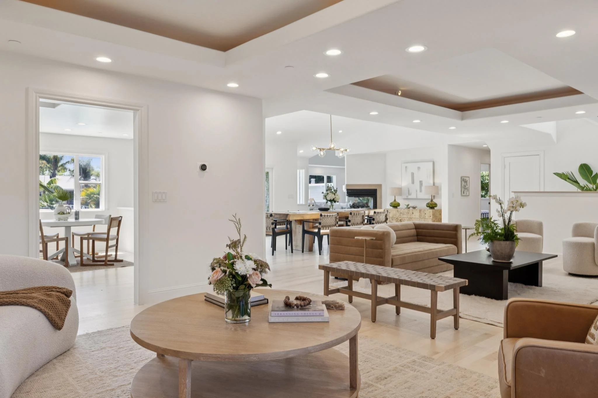 Open concept living room and dining area with white walls, recessed lighting, and wooden ceiling accents. Features a beige sofa, armchairs, a black coffee table, and decorative plants and flowers.