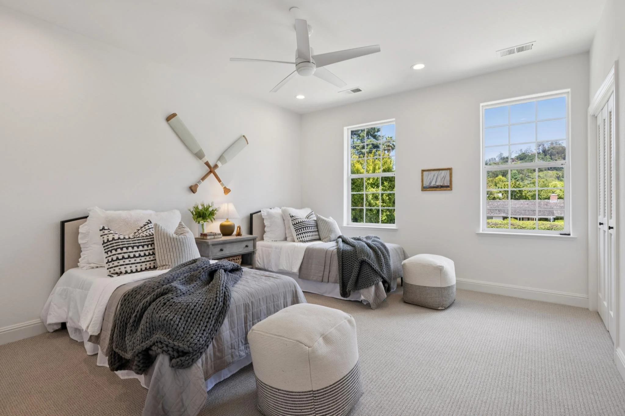 Bright bedroom with two beds, white walls, large windows showing greenery, ceiling fan, and decor including paddles and framed art.