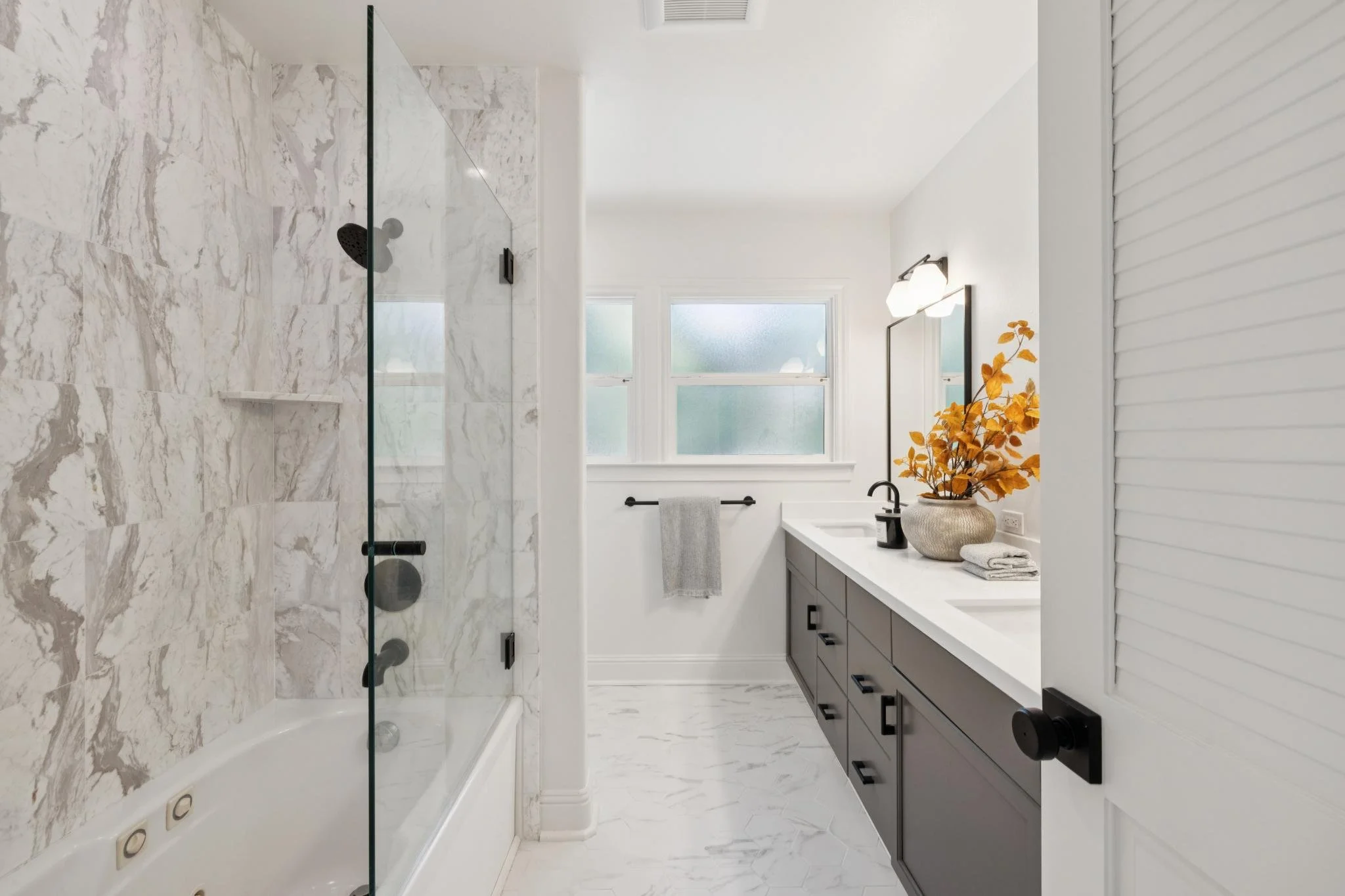 Modern bathroom with a marble shower on the left, a large window with frosted glass in the center, and a dark vanity with a white countertop on the right, decorated with a vase of orange leaves and towels.