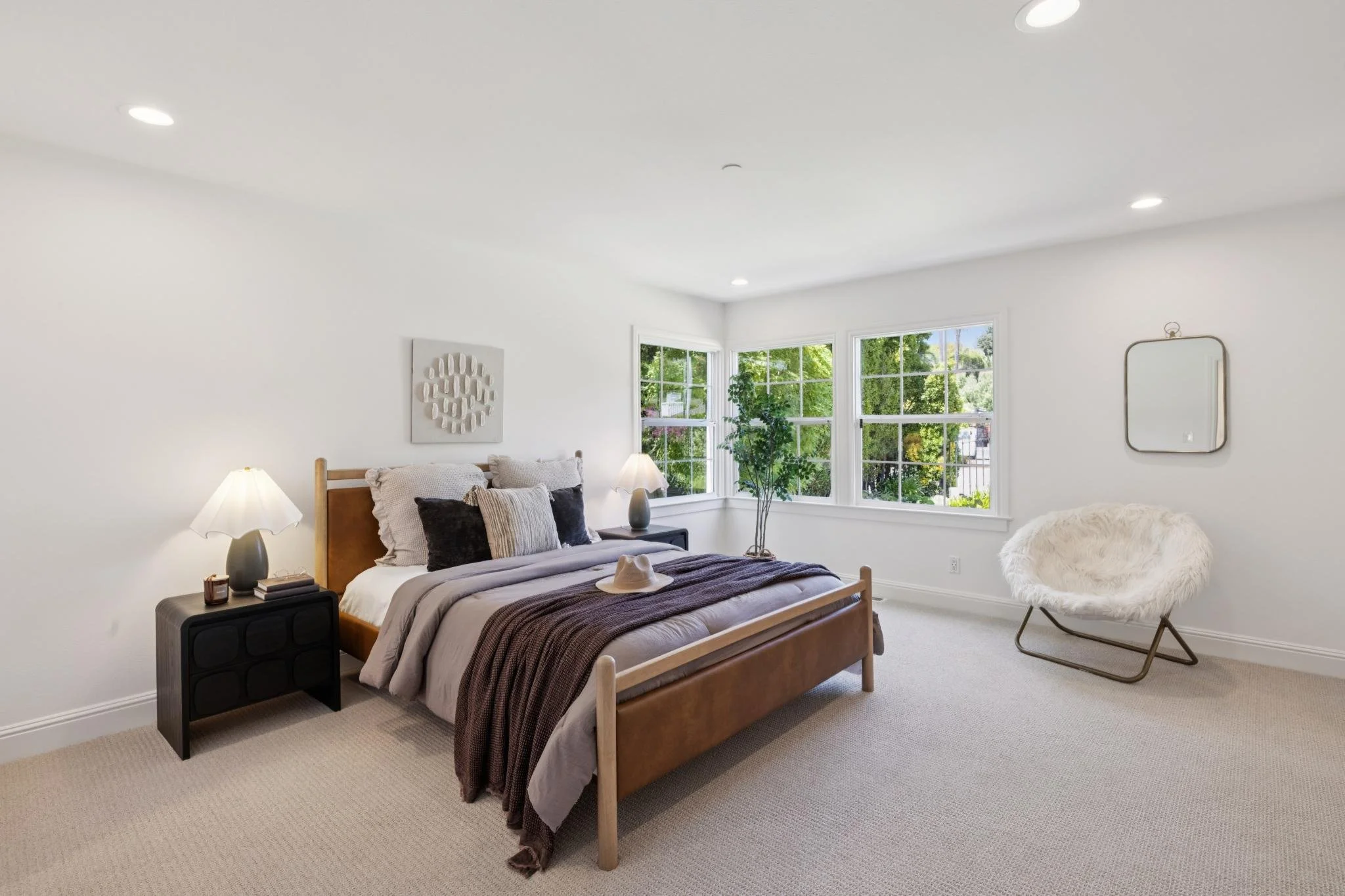 A bedroom with white walls and a large window showing green trees outside. The bed has a wooden frame with beige and dark pillows, a beige blanket, and a hat on top. There are two nightstands with lamps, one with books. Next to the window, there is a