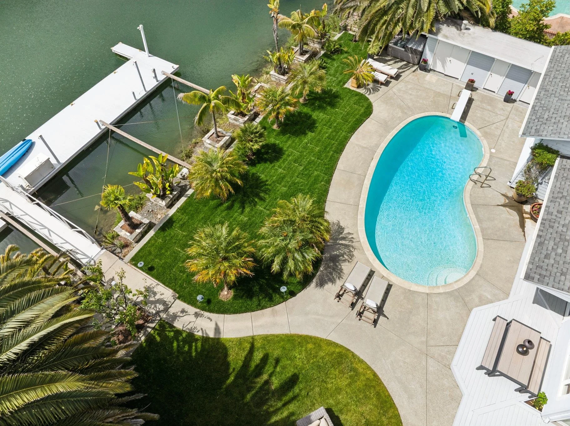 Aerial view of a backyard with a kidney-shaped swimming pool, surrounded by a concrete deck, sun loungers, a small grassy area with a few trees, and a waterway with a dock and canal boats. There is also a white shed and tropical plants.