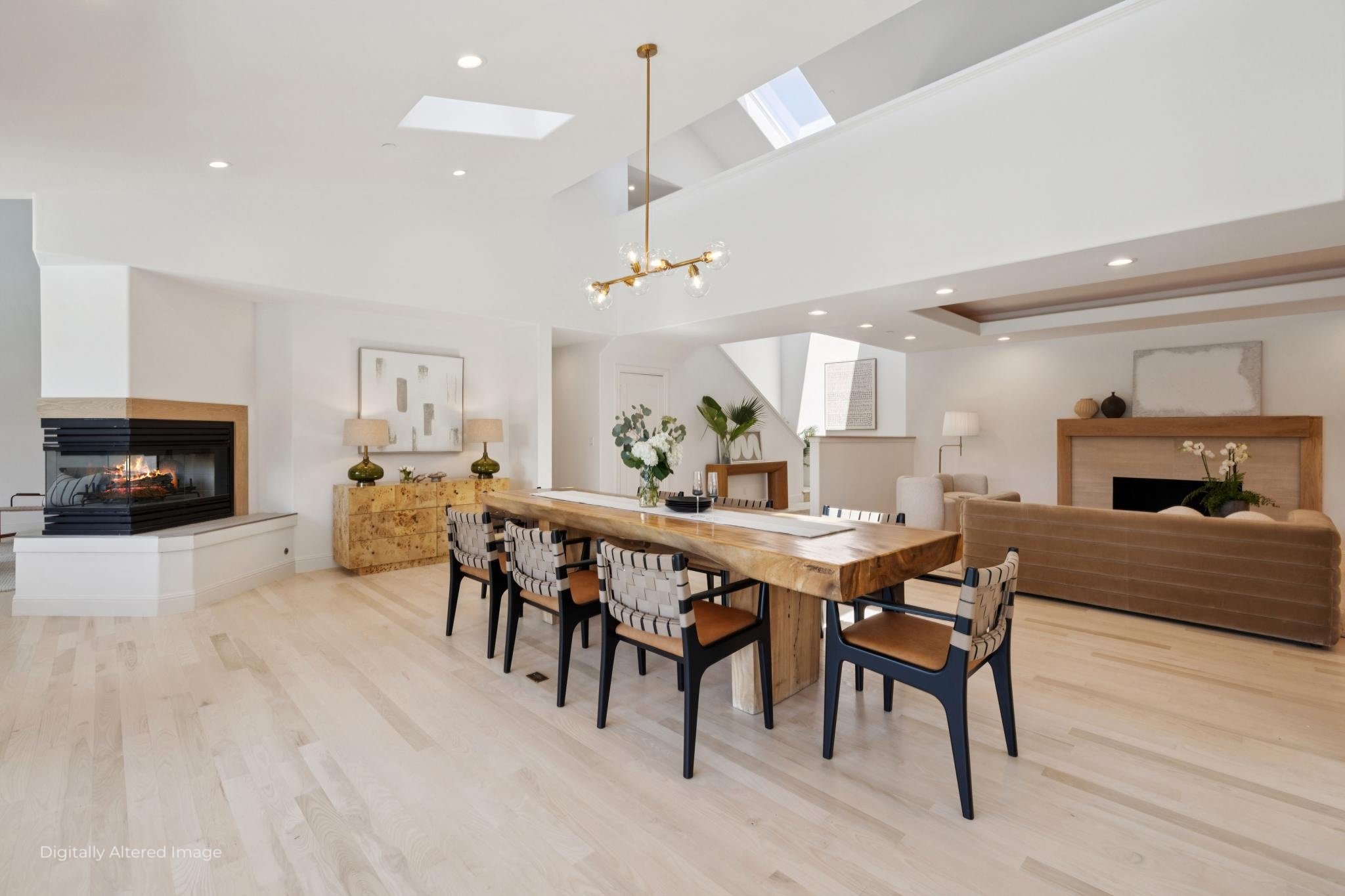 Living room with a wooden dining table, black and striped chairs, a beige sofa, modern fireplace, decorative vases, plants, artwork, and skylights in a bright, minimalist style.