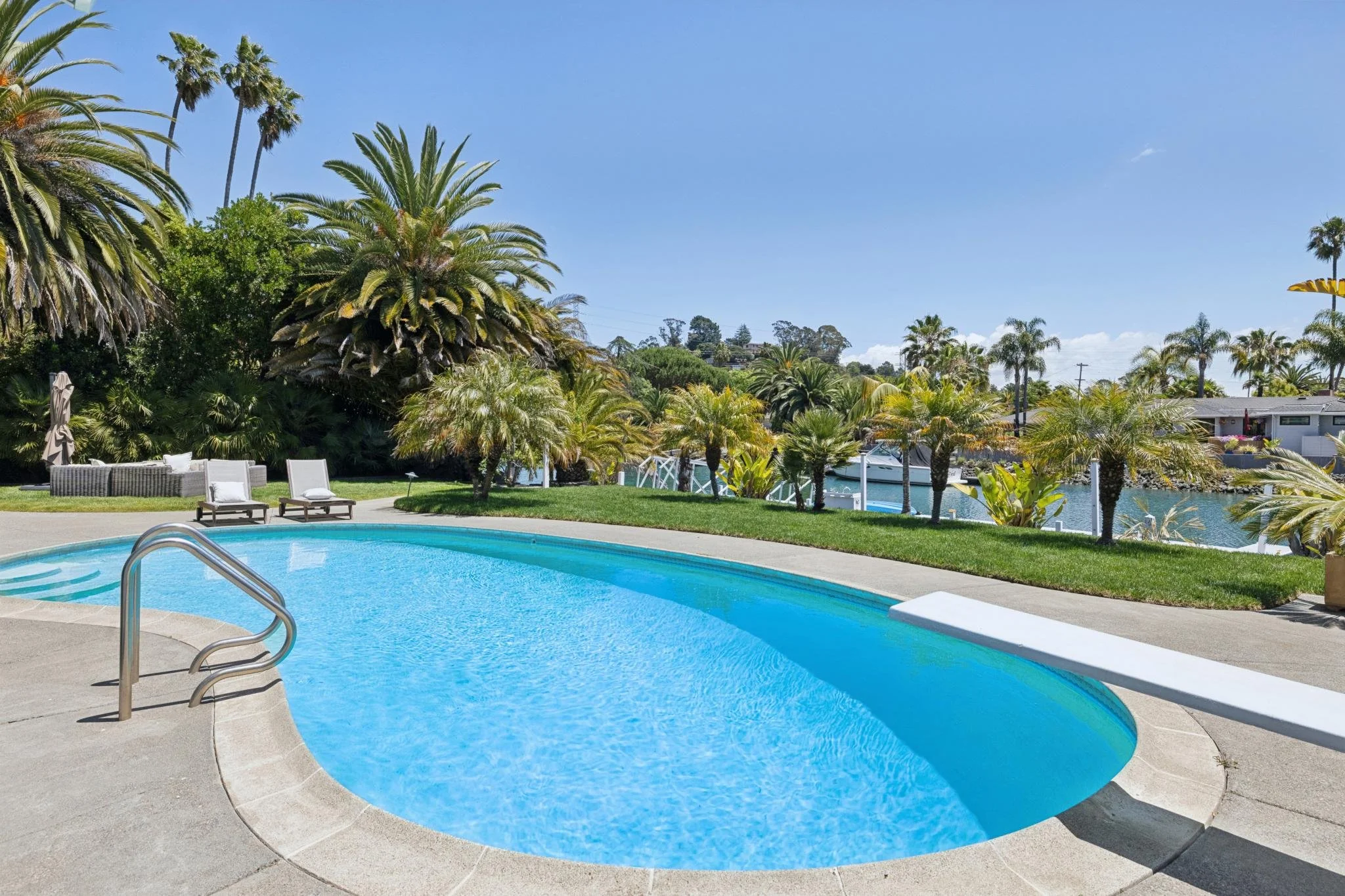 An outdoor swimming pool with a curved shape, surrounded by a concrete deck, and poolside lounge chairs. There are palm trees and lush greenery in the background under a clear blue sky.