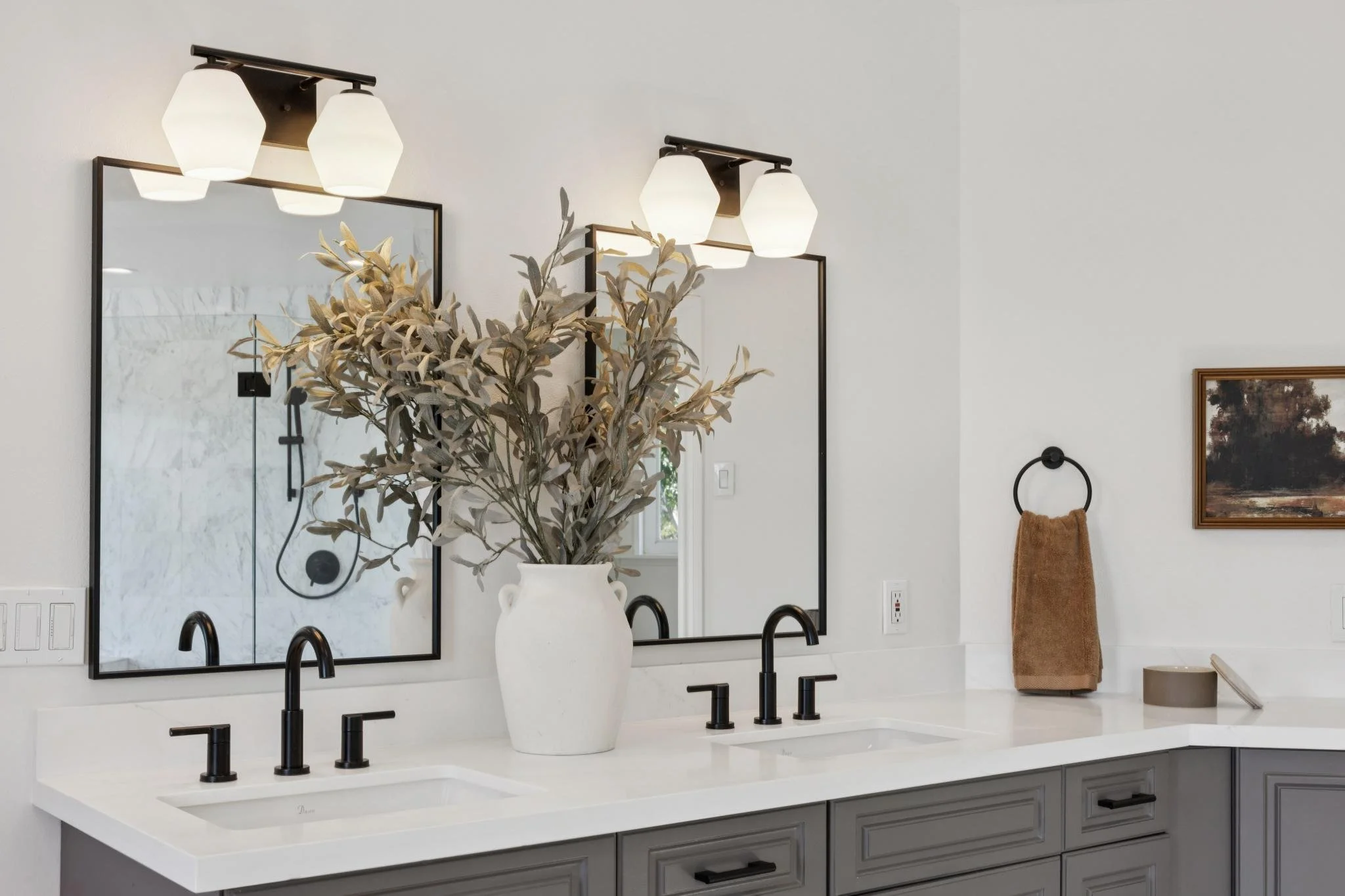 Modern bathroom with double vanity, black faucets, large mirror, potted plant, and wall-mounted light fixtures.