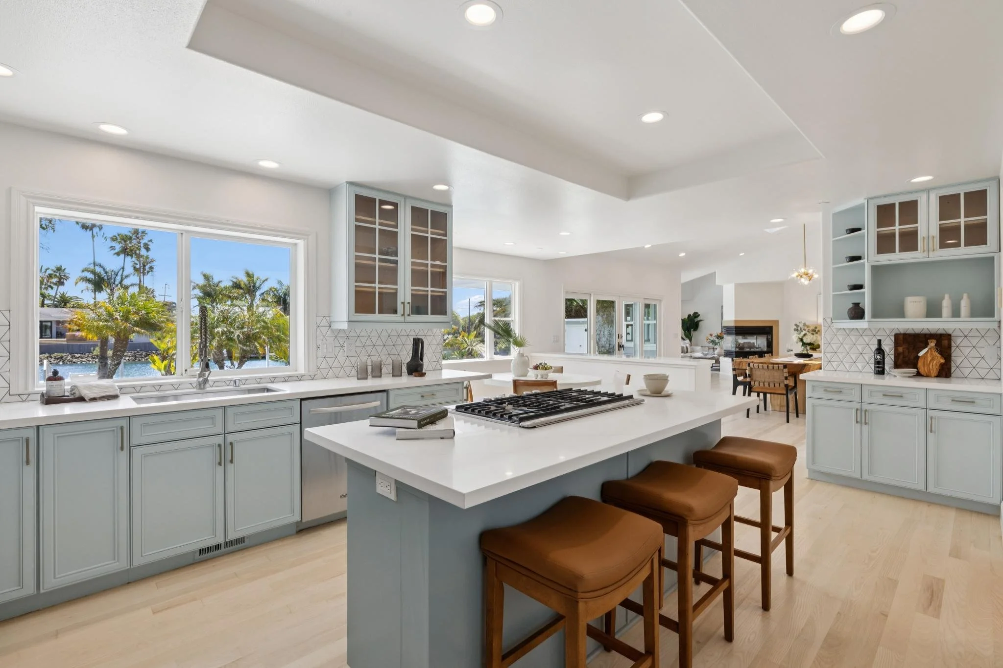 Bright, modern kitchen with white cabinets, large windows showing an outdoor view of palm trees, an island with brown stools, and open living space in the background.