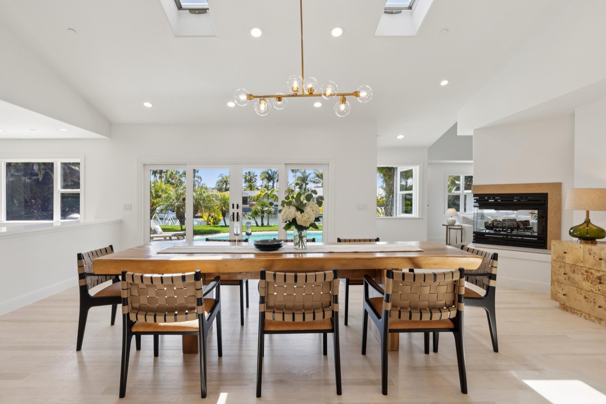 A modern dining room with a wooden table surrounded by chairs, floral centerpiece, large windows with a view of green trees, and a ceiling with skylights and a chandelier.