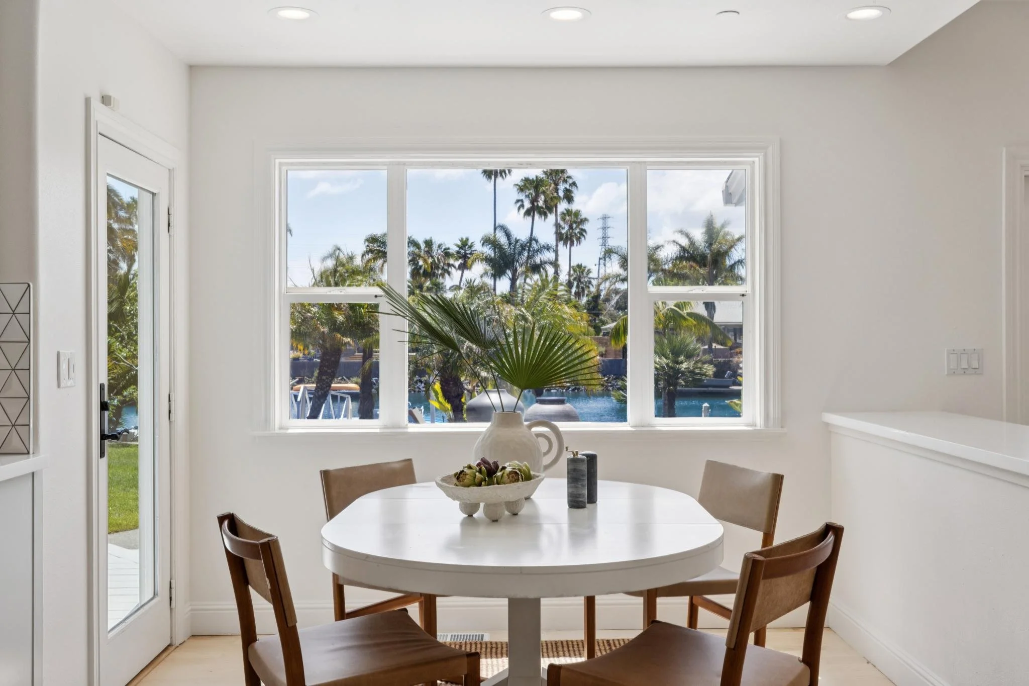 A bright room with large window overlooking tropical trees and water, a white round table with a decorative bowl of artichokes and other items, surrounded by wooden chairs.