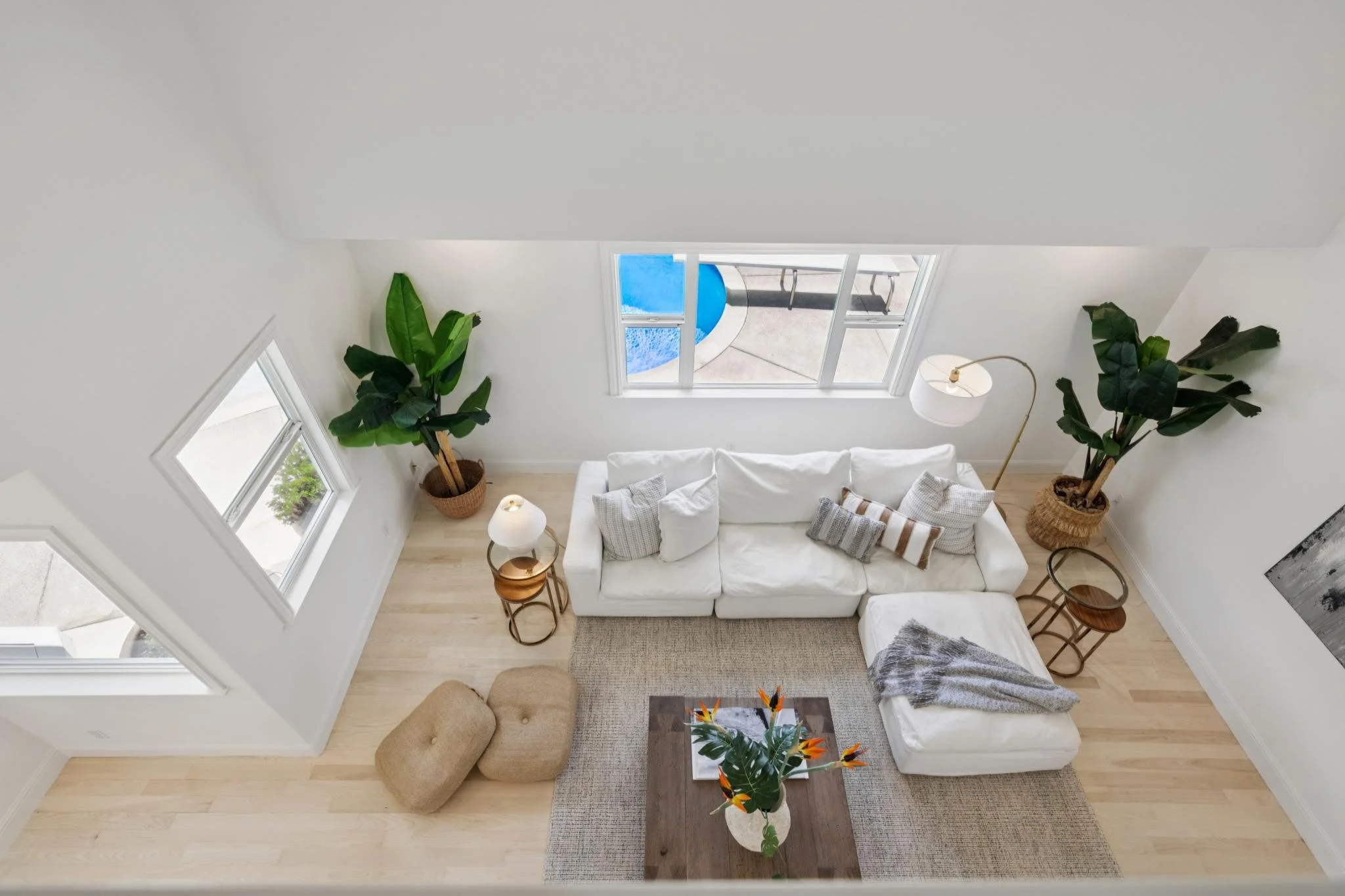 Living room with white sofa, beige ottomans, wooden coffee table with a floral arrangement, large green potted plants, side tables, a floor lamp, and windows showing a view outside.