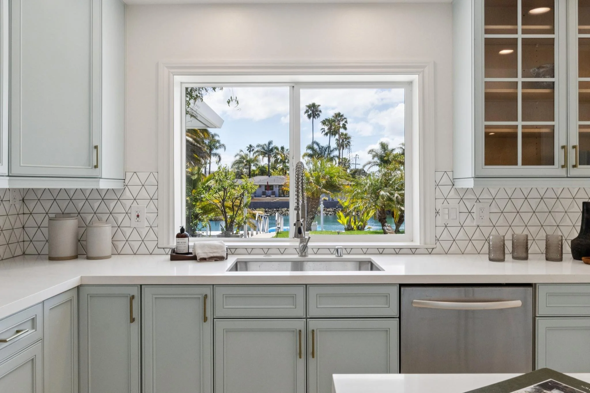 Kitchen with a large window showing a view of palm trees, blue sky with clouds, and nearby houses.