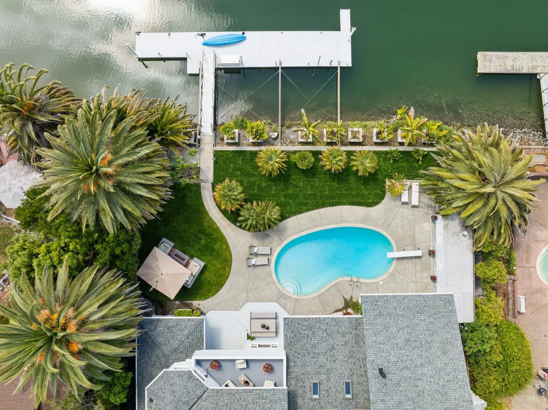 Aerial view of a backyard with a swimming pool, palm trees, a green lawn, a patio with outdoor furniture, a dock on the water with a kayak, and neighboring houses.