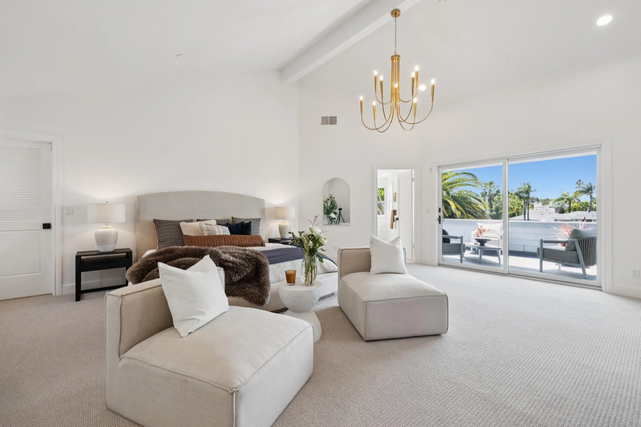 Bright living room with white walls, large sliding glass doors opening to a balcony with outdoor seating, beige carpet, white and brown furniture, and a gold chandelier.