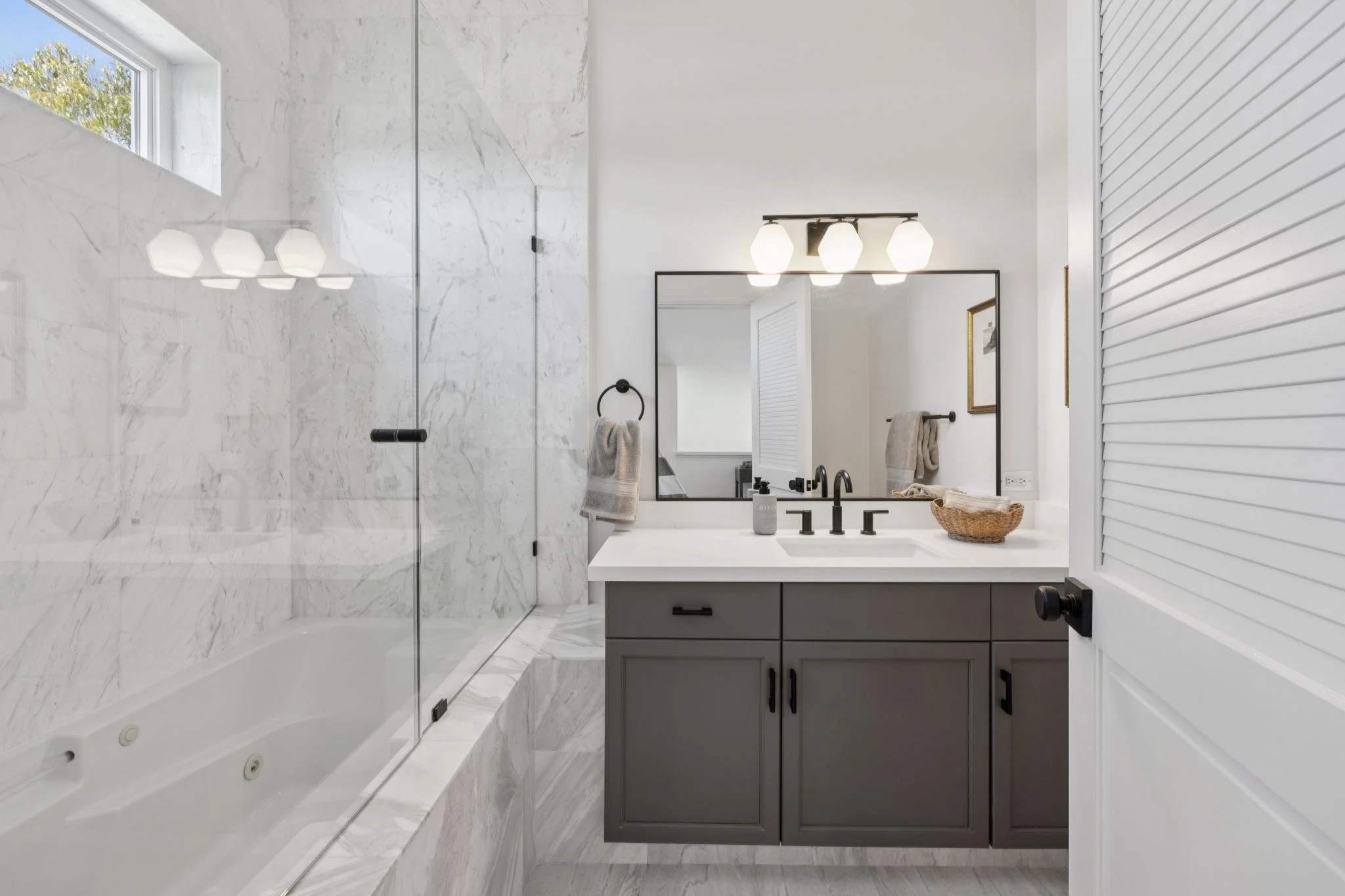 Modern bathroom with white marble walls, a large mirror above a gray vanity with black hardware, black fixtures, and a basket on the counter. There is a glass-enclosed shower to the left with a built-in bench and a window above, allowing natural ligh