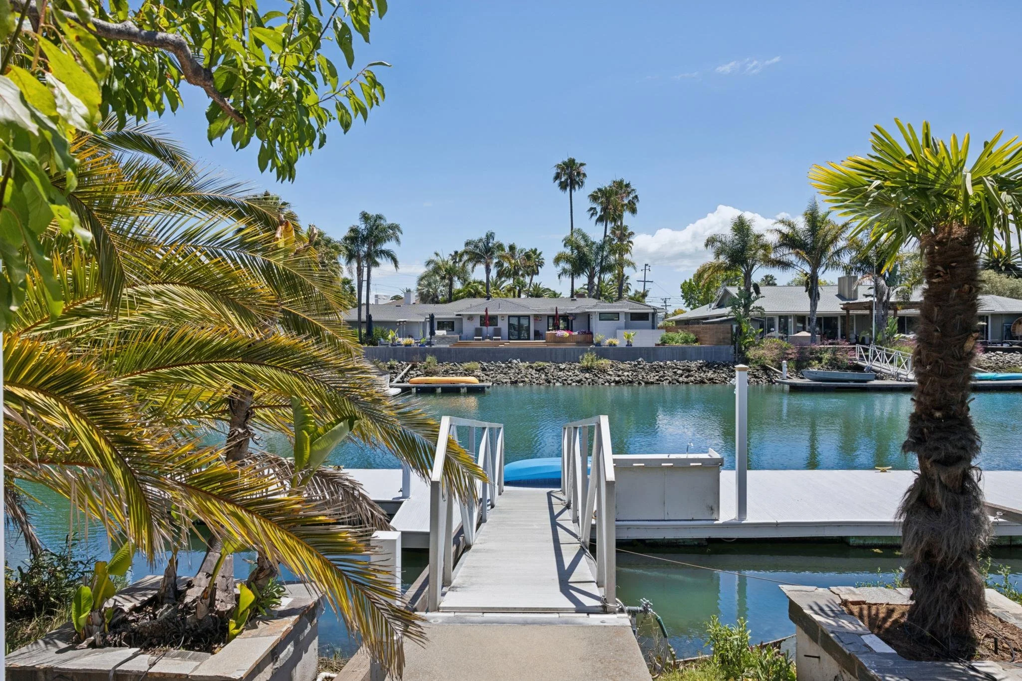 A waterfront view with palm trees, a dock, and houses across the water under a blue sky.