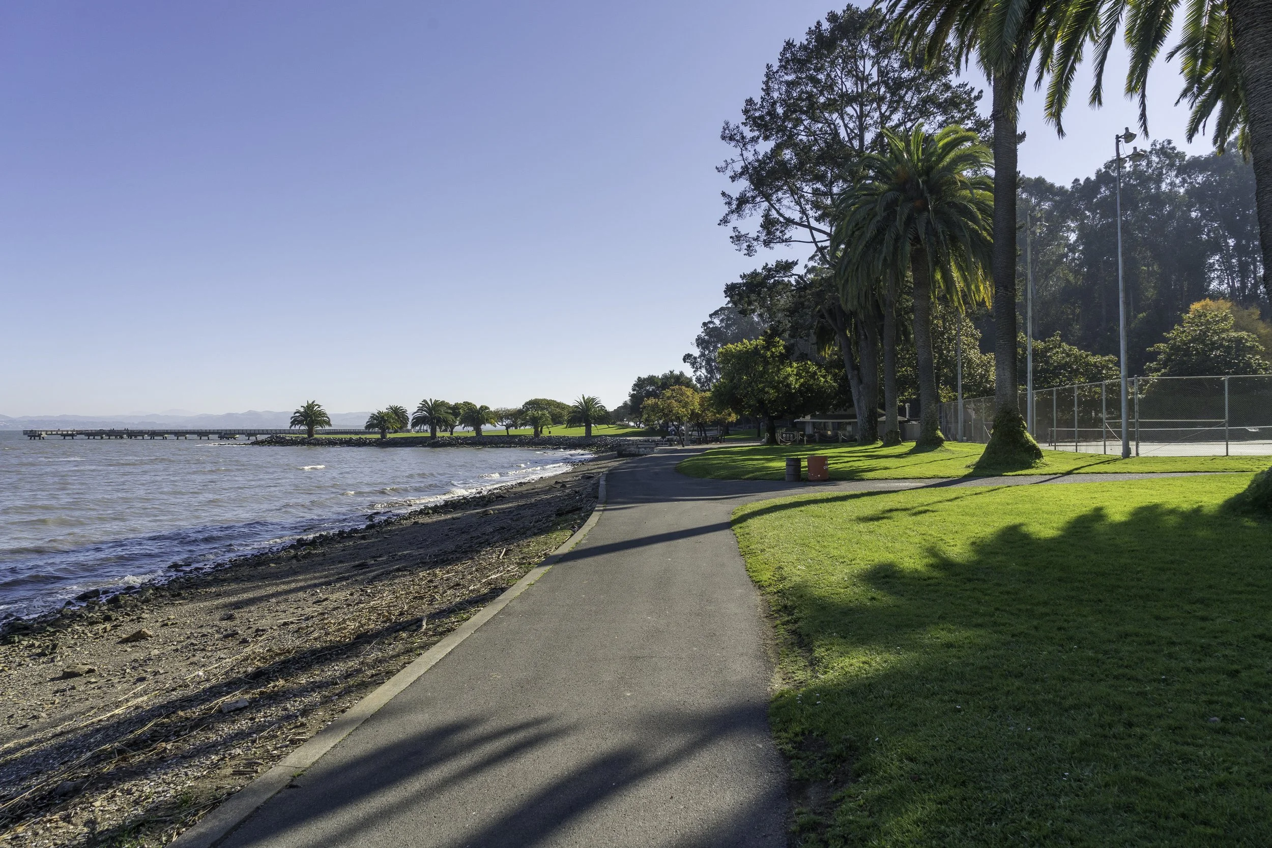 A lakeside park with a paved walking trail, grassy area, tall palm trees and light poles. The water has gentle waves and a pier extends into the lake, with mountains visible in the background. Clear blue sky overhead.
