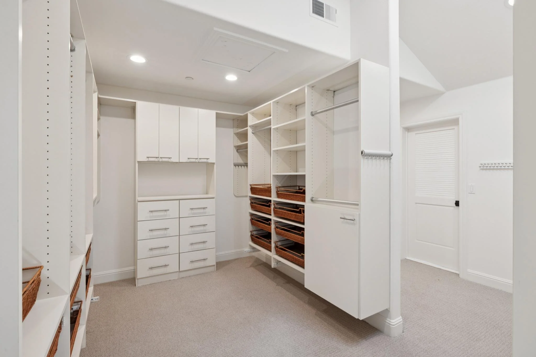 A walk-in closet with white built-in shelves, drawers, and baskets for storage, illuminated by ceiling lights.