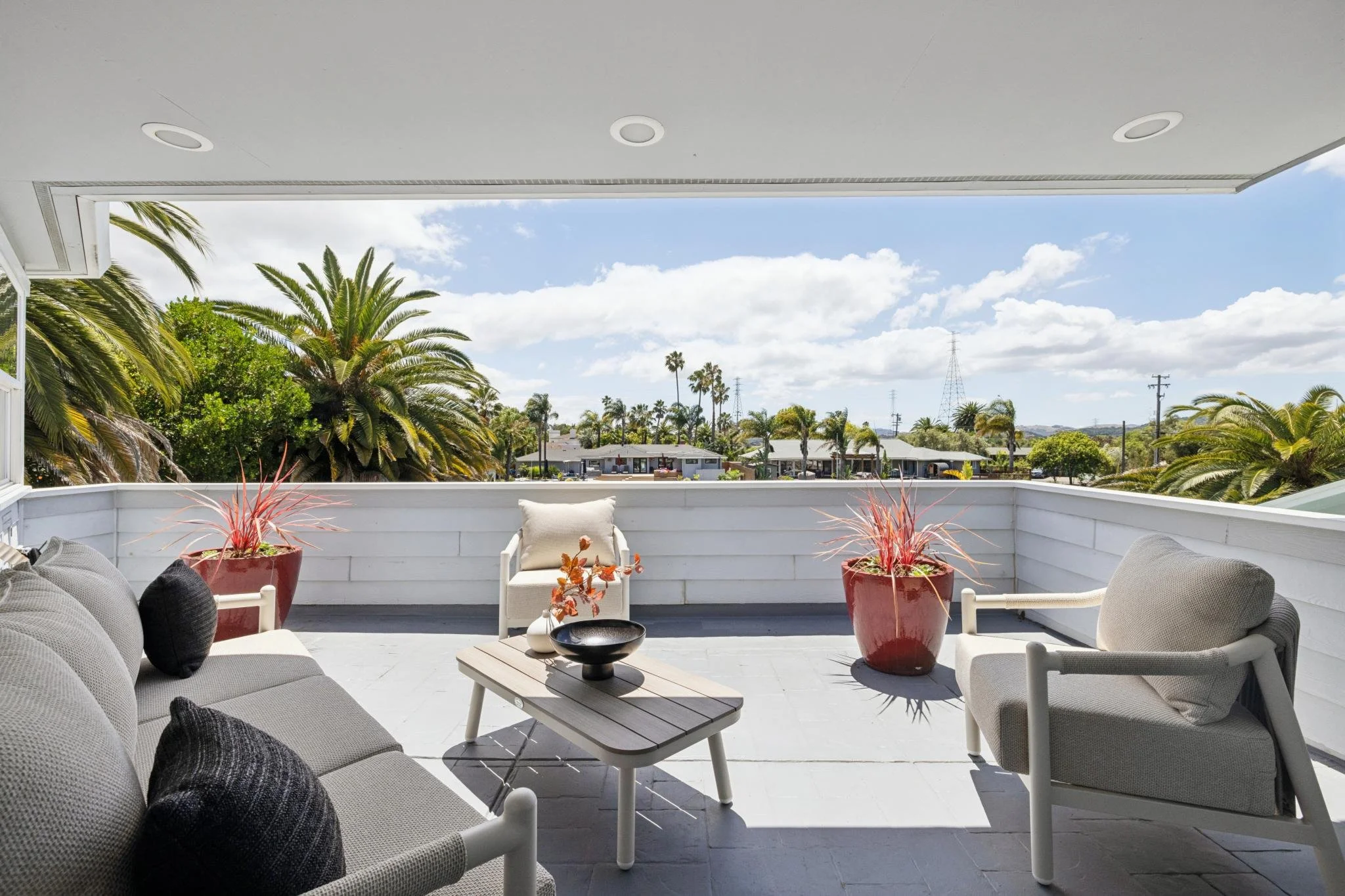 A balcony with outdoor furniture, including a gray cushioned sofa with black pillows, a white armchair with a beige pillow, a small table with a black bowl, and two large red pots with red plants. The balcony has a white railing and overlooks a neigh