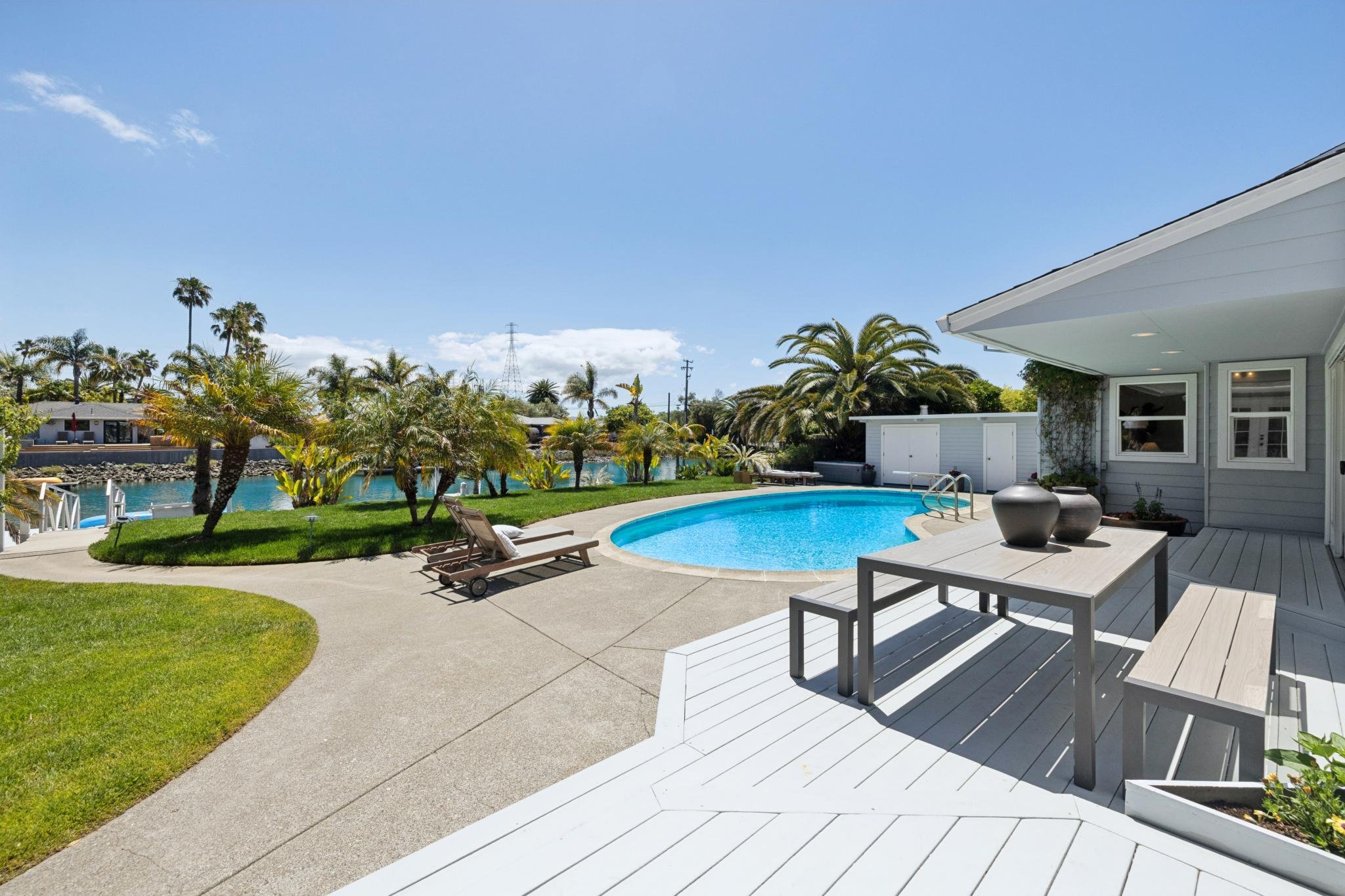 View of a backyard with a swimming pool, palm trees, and a patio with outdoor furniture under a clear blue sky.