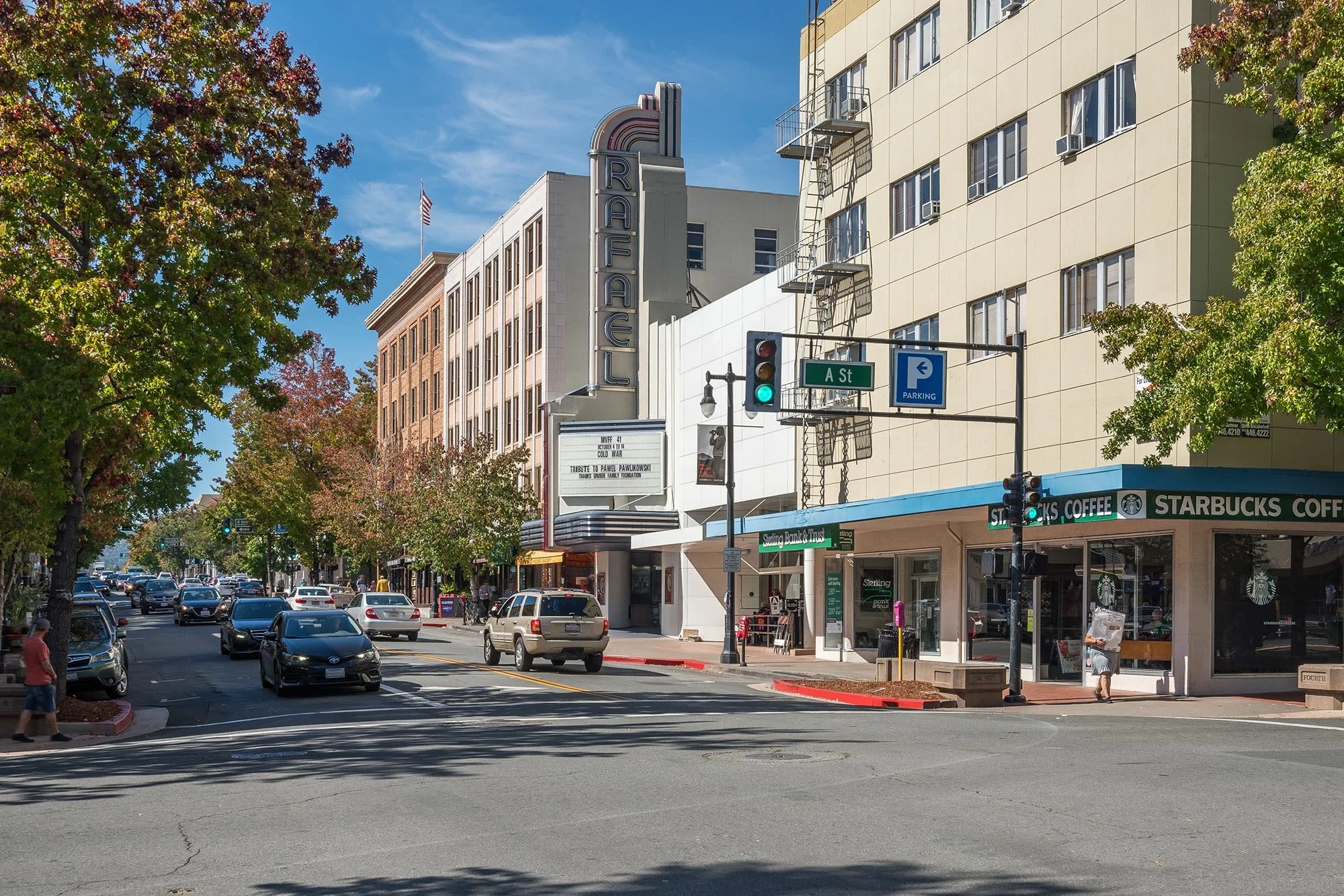 Downtown street scene with cars, trees, a building with a large vertical sign that reads 'Rafaels,' a Starbucks coffee shop, street signs for A Street and parking, traffic lights, and pedestrians.