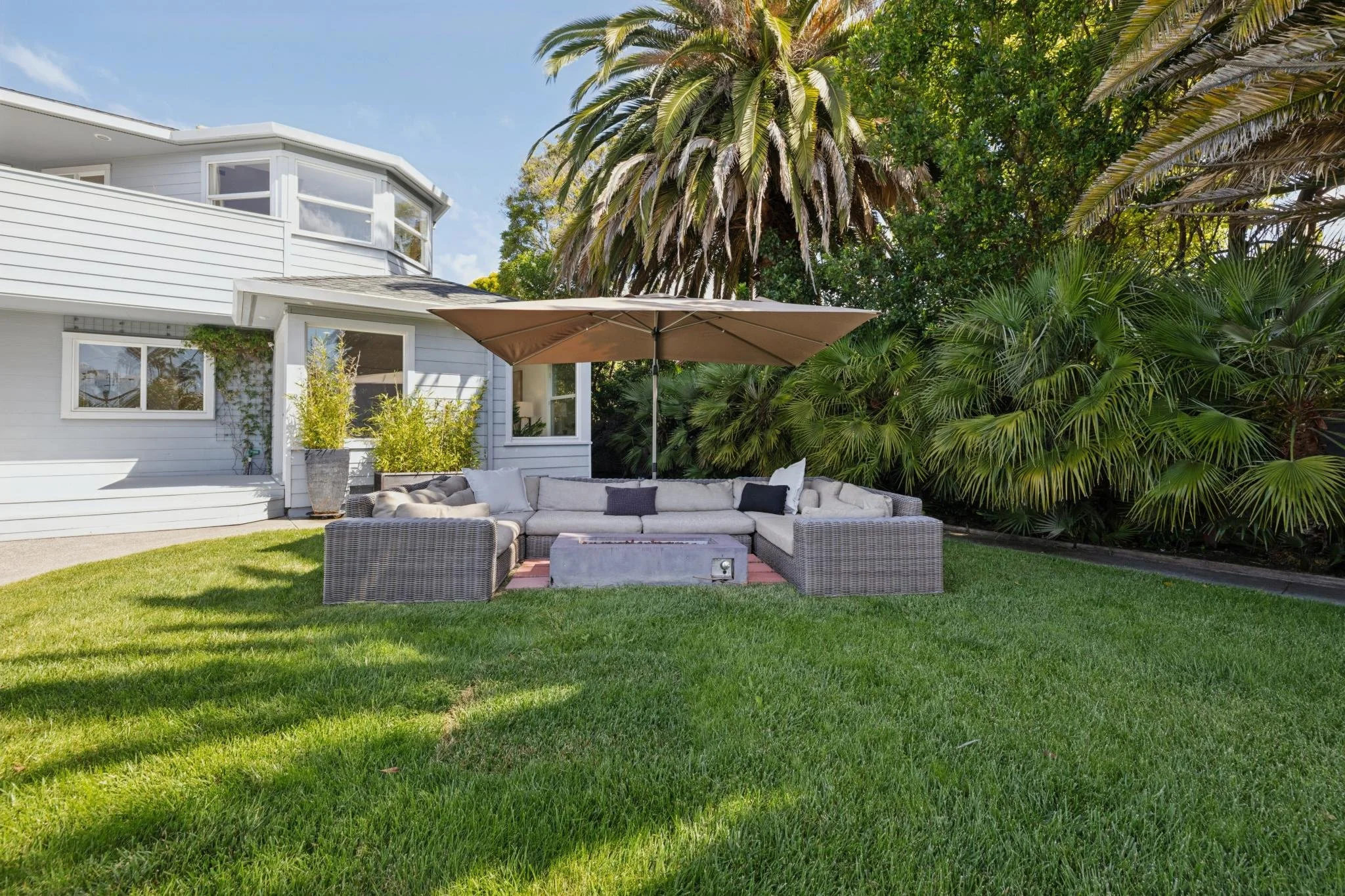 A backyard patio area with gray wicker outdoor furniture, cushions, and a large beige umbrella. The area is surrounded by green grass and lush, tropical plants, with a white house in the background and tall palm trees overhead.