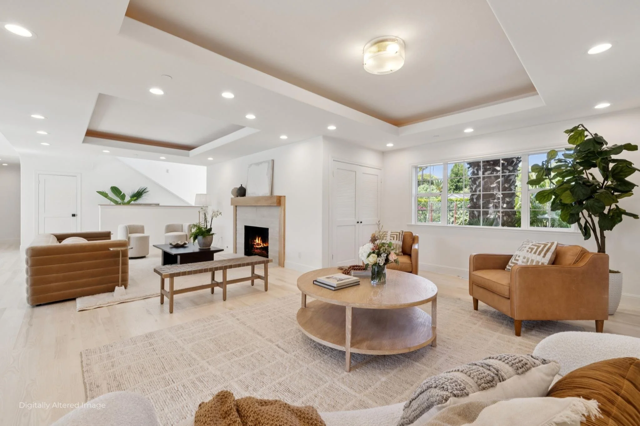 Living room with white walls, large window showing greenery outside, beige sofas, coffee table with books and flower arrangement, fireplace, decorative vases, and potted plant.