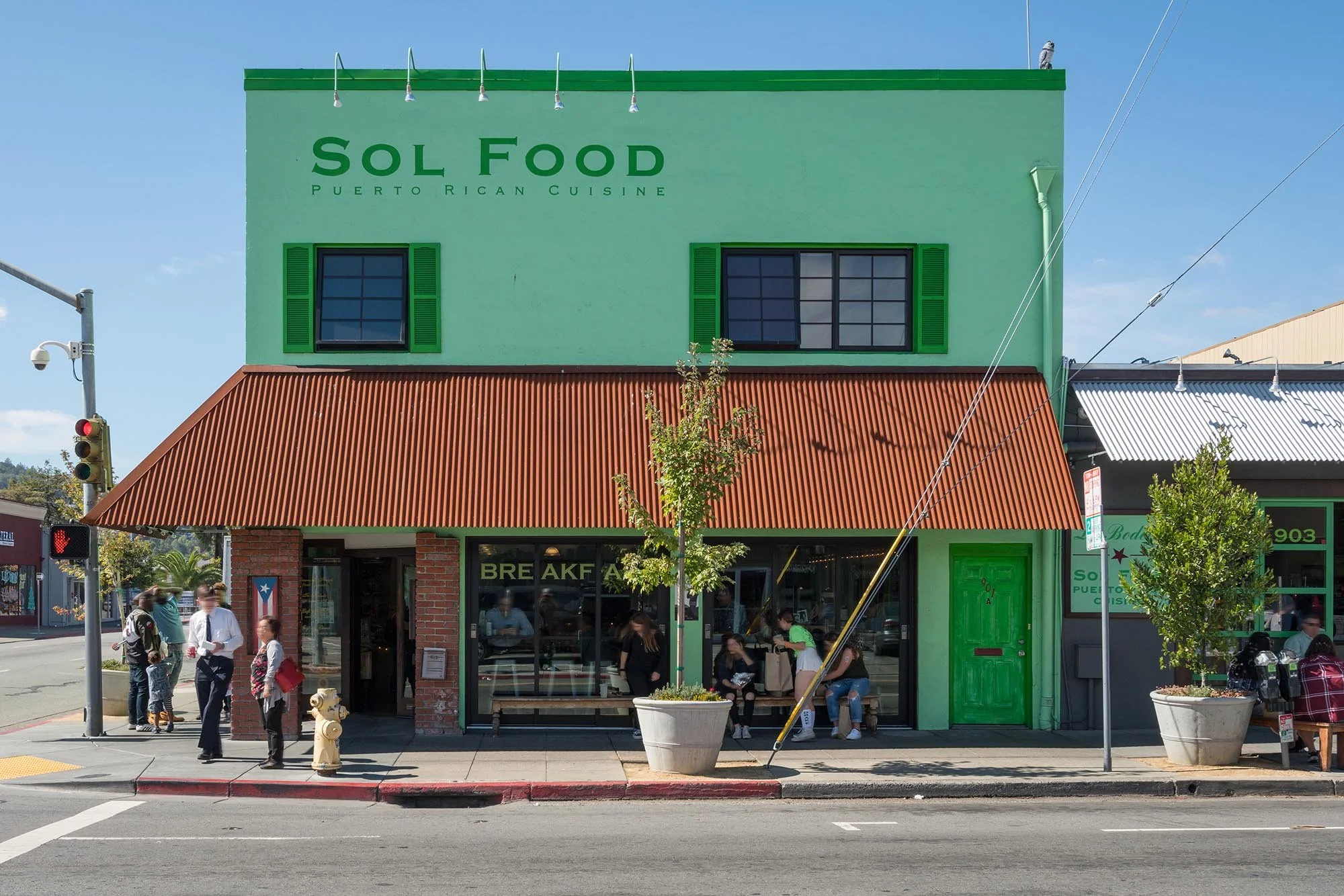 A colorful building with green walls, green window shutters, and a red metal roof. The sign reads 'Sol Food Puerto Rican Cuisine.' There are people walking and sitting outside, and a traffic light is visible on the street corner.