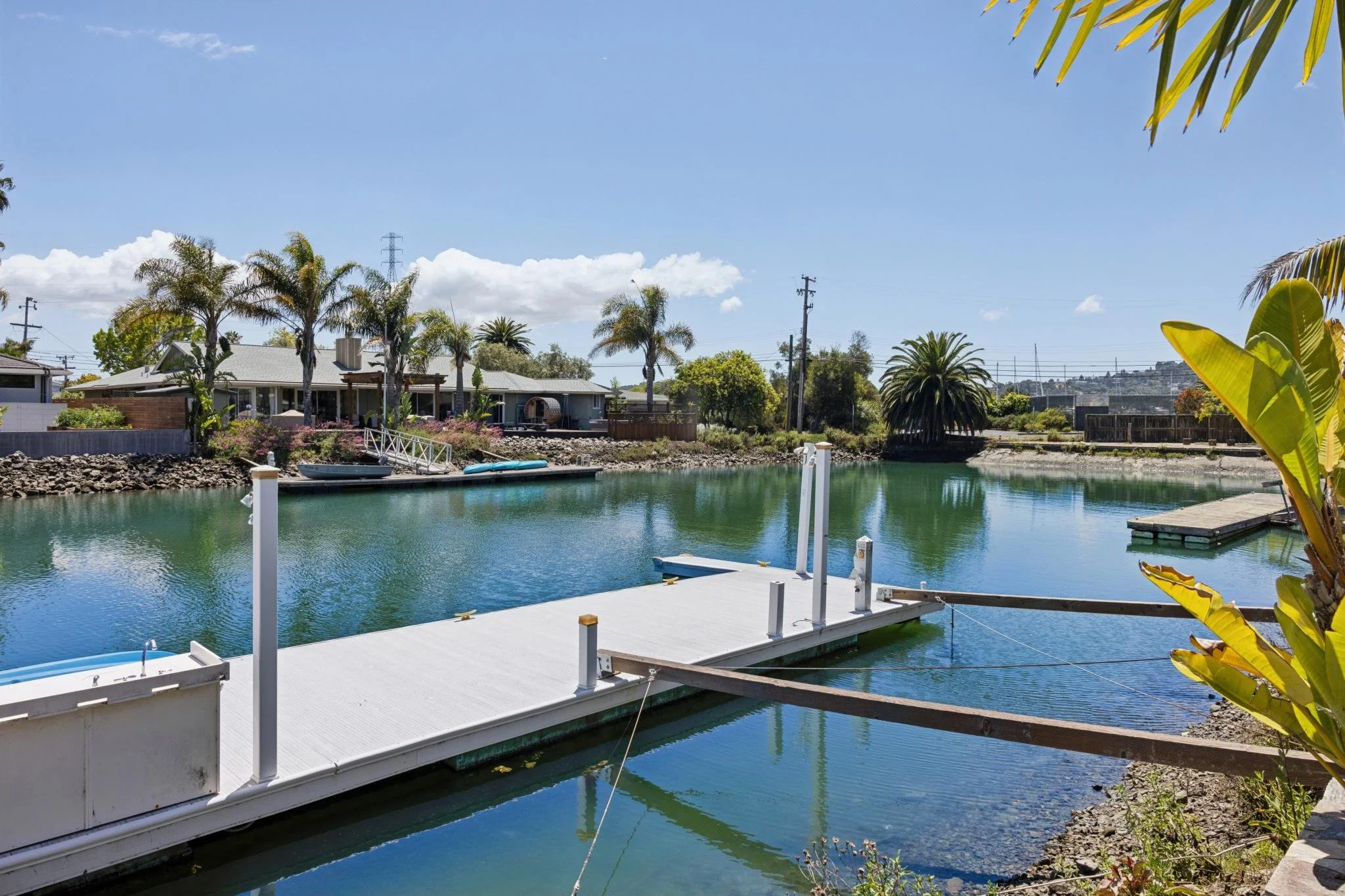 Residential backyard with a canal, a dock, boats, palm trees, and a house under a blue sky with some clouds.