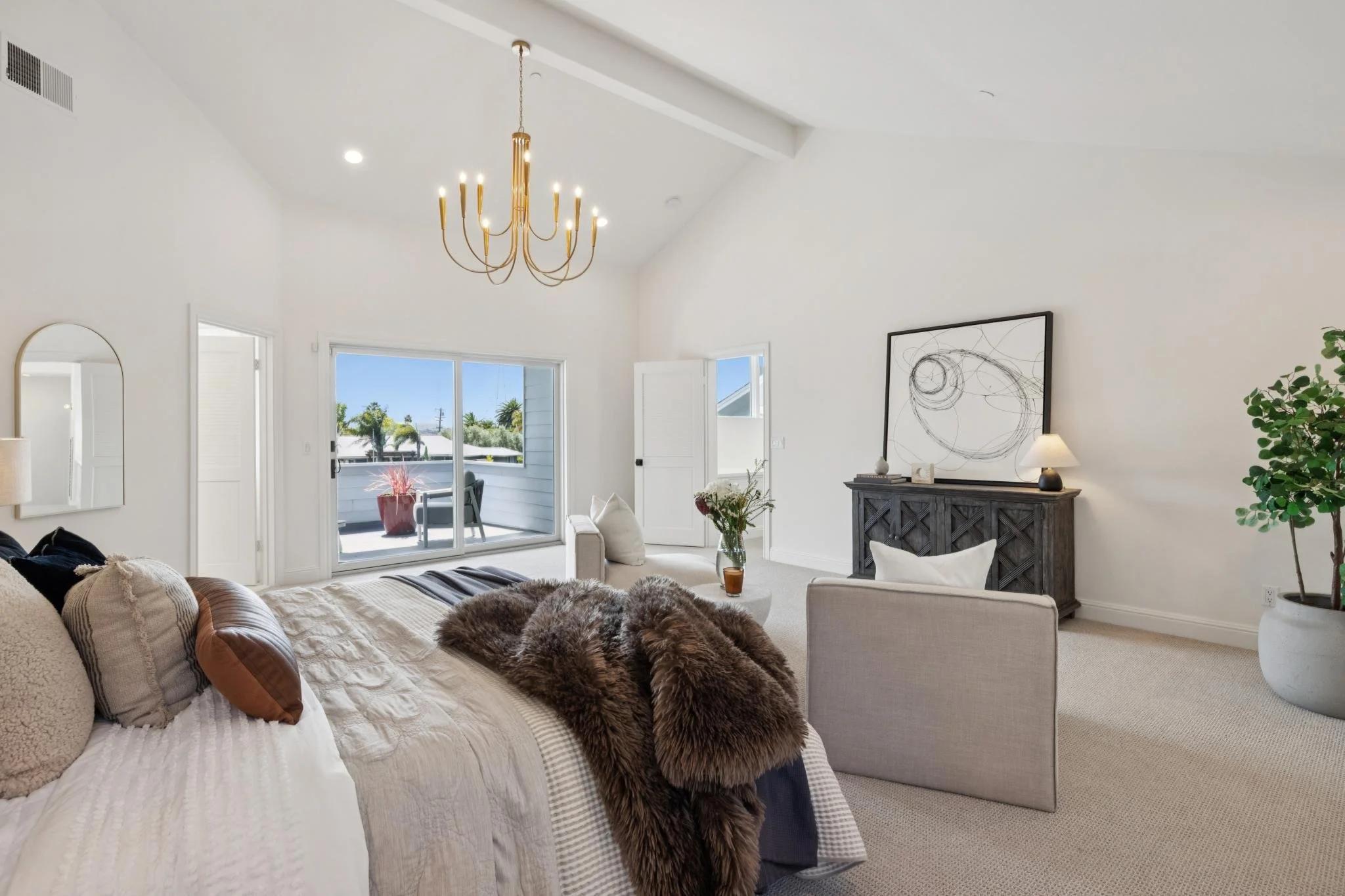 Living room with white walls, a beige sofa with pillows, a faux fur throw, a side table with a plant and a glass of drink, a black console with wall art on top, a lamp, a tall green plant, a ceiling chandelier, and sliding glass doors leading to an o