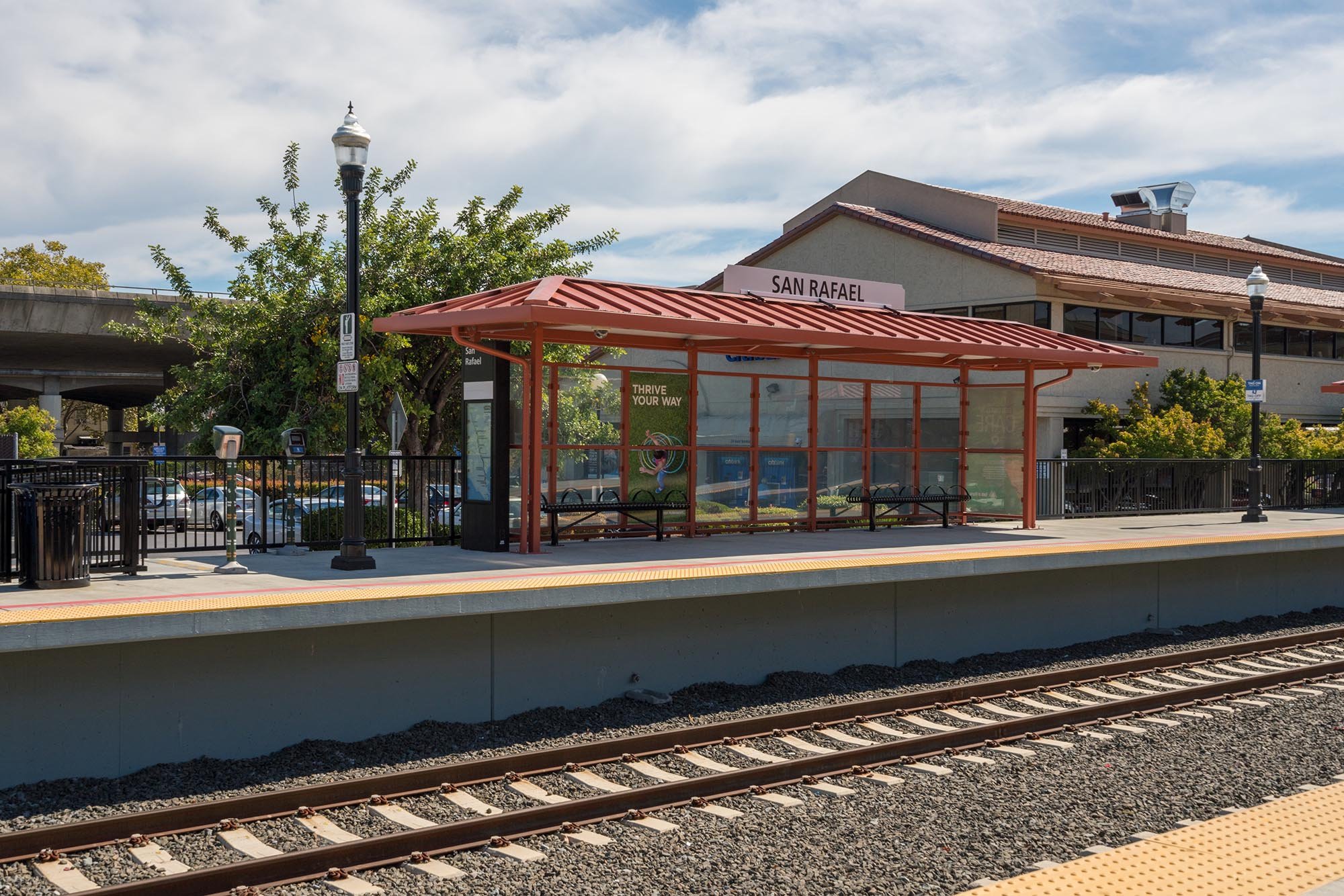 Train platform with a shelter, benches, trees, a streetlamp, and a building labeled 'San Rafael' in the background. Railroad tracks are visible in the foreground.
