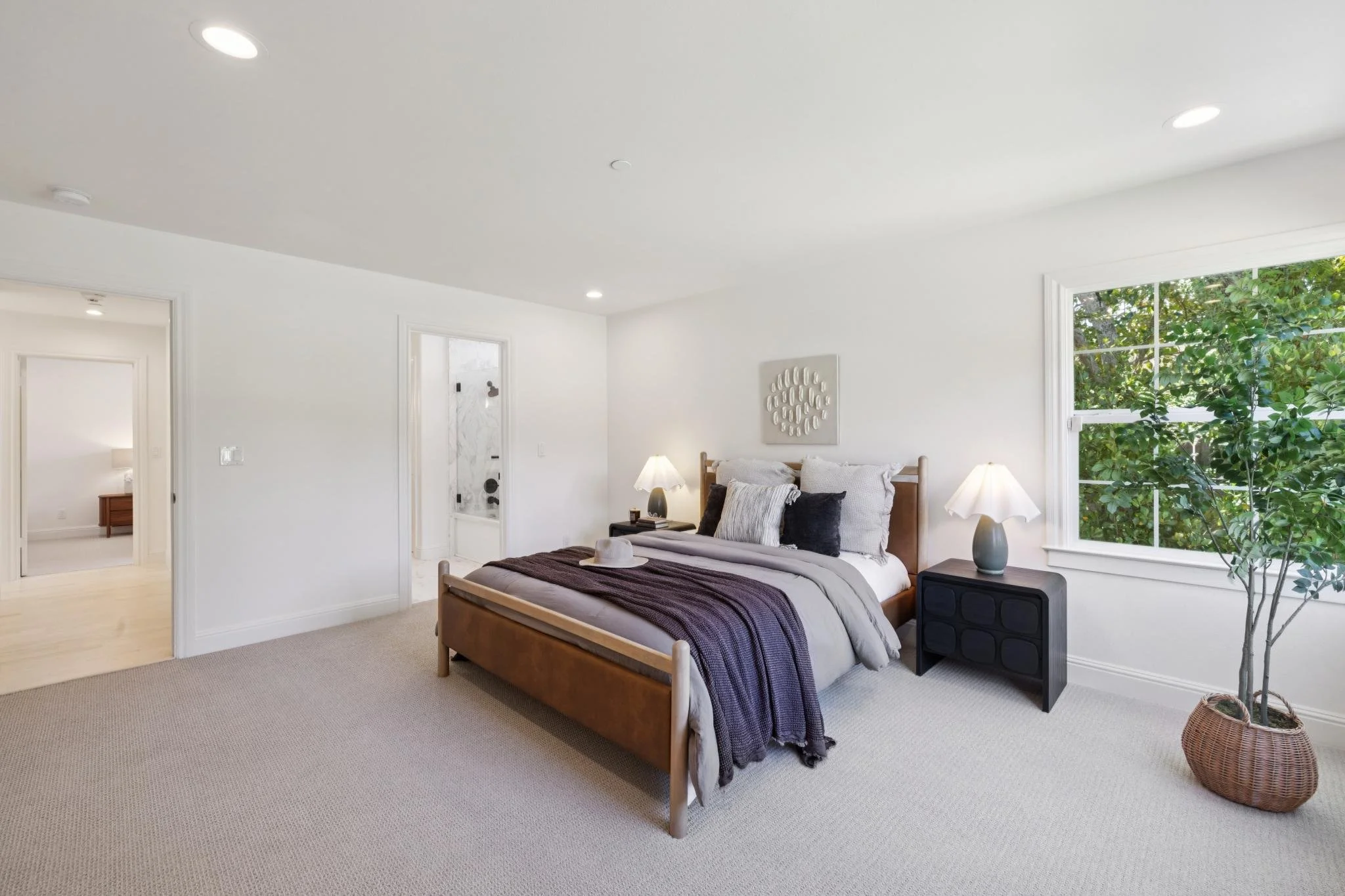 A bedroom with a wooden bed frame, white bedding, and dark pillows, illuminated by two table lamps, with a large window revealing green foliage outside.