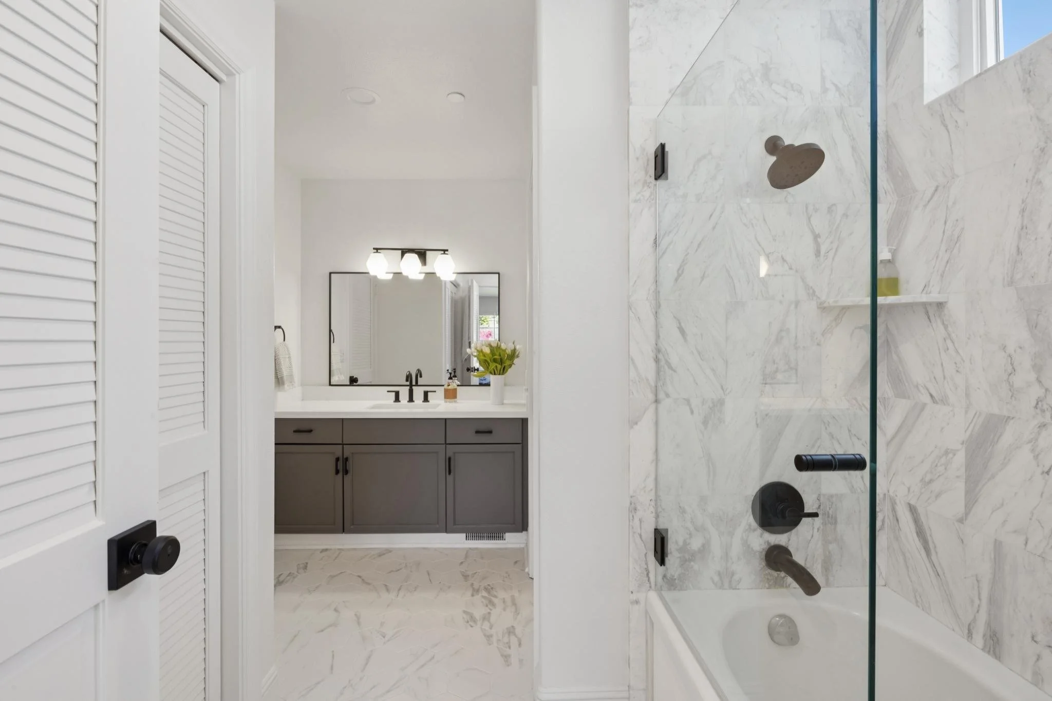 Modern bathroom with white marble walls and floor, gray vanity with black fixtures, a mirror, and a glass-enclosed shower with a black showerhead.