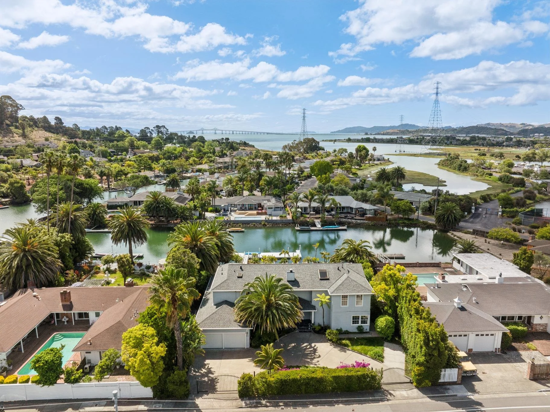 Aerial view of a residential neighborhood with houses, palm trees, a waterway, and a bridge in the distance under a partly cloudy sky.