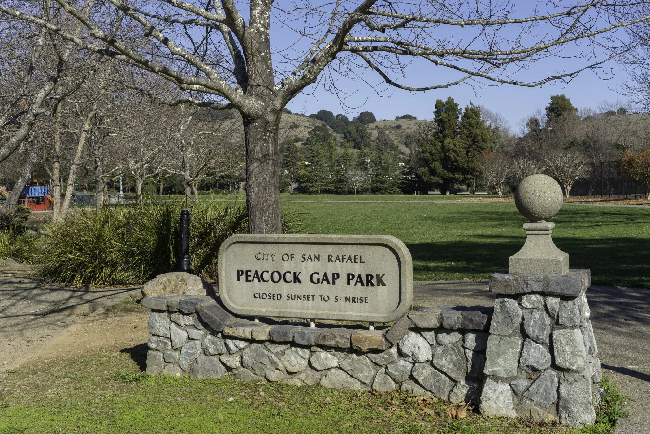 Sign at Peacock Gap Park in San Rafael stating it is closed from sunset to sunrise, with trees and grass in the background under a clear blue sky.