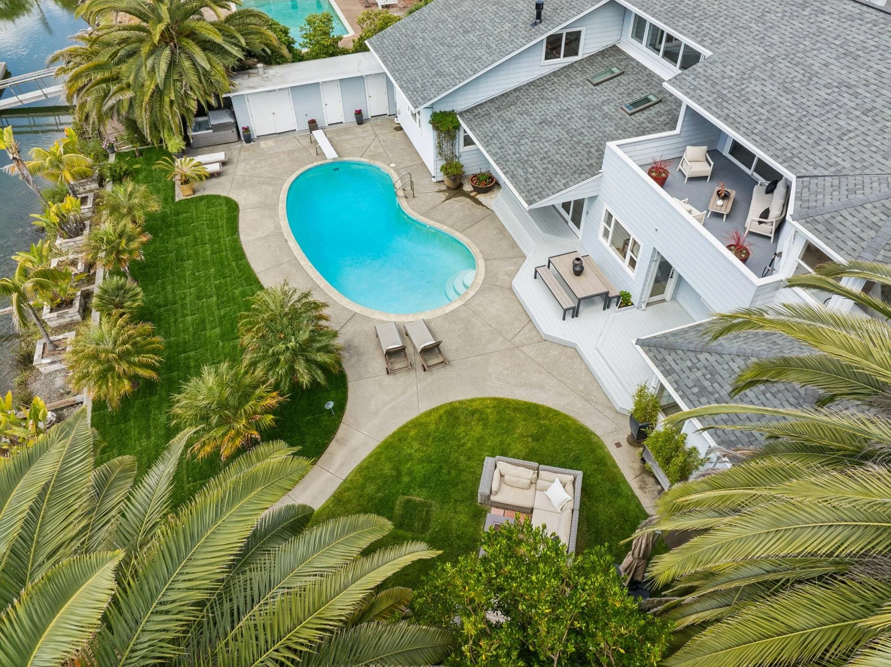 Aerial view of a backyard with a kidney-shaped swimming pool, lush green grass, tropical palm trees, patio furniture, and a white house with a second-story balcony.