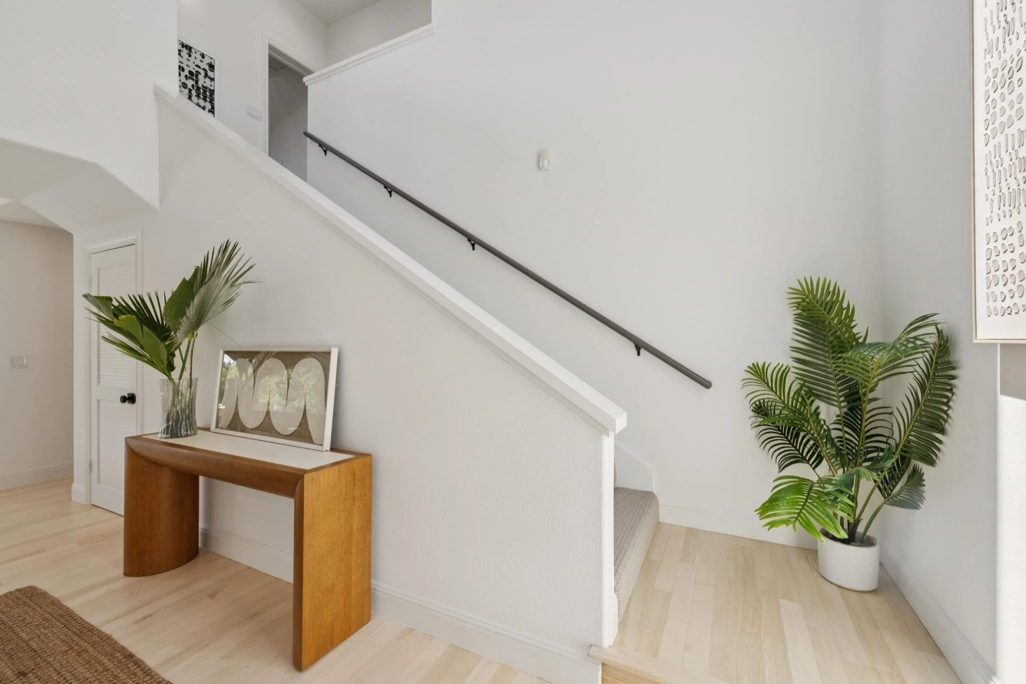 Interior of a modern house with light-colored hardwood floors, a white wall with a staircase, some green plants in pots, and a wooden table with framed artwork and a glass vase on top.