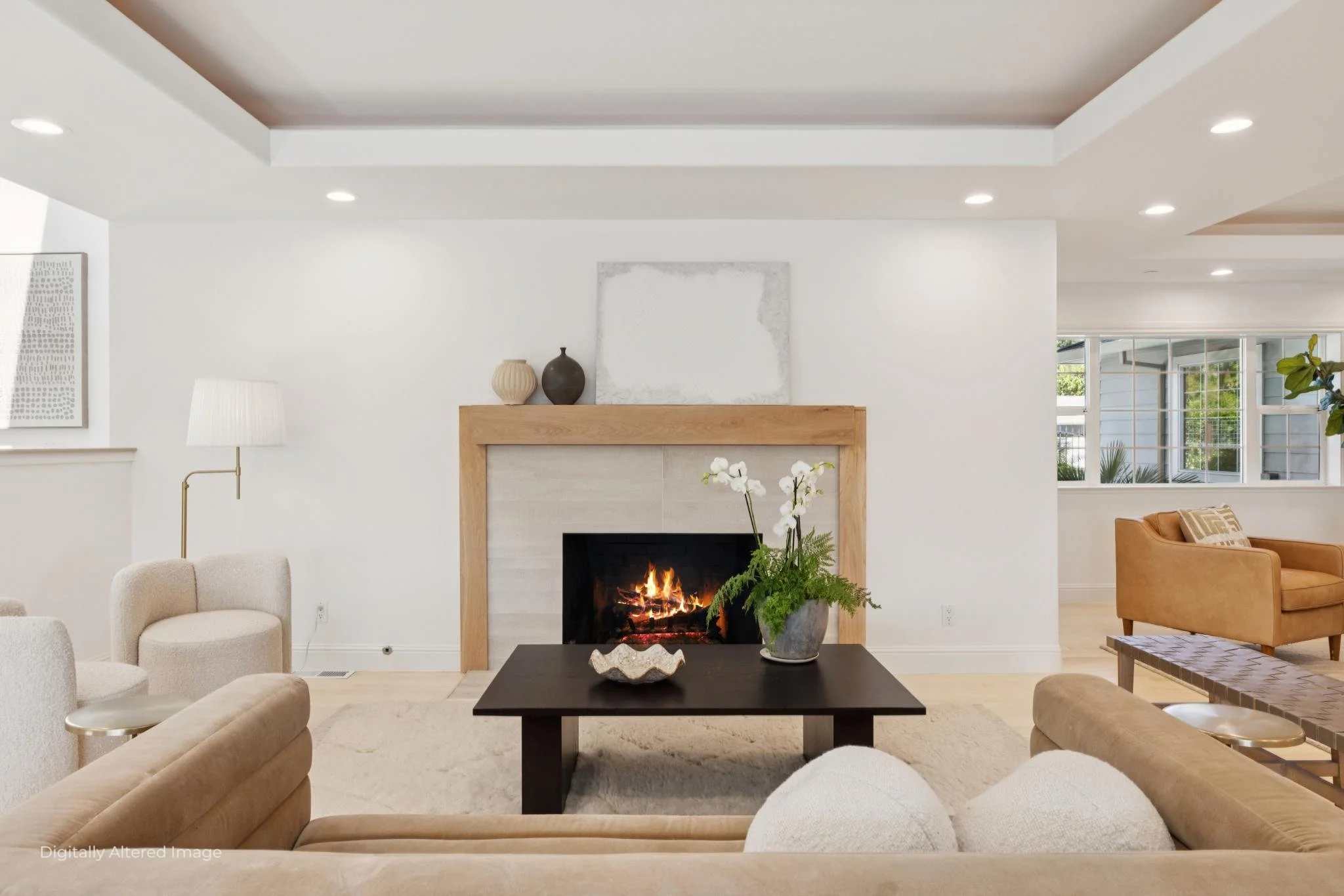 A living room with a fireplace, beige sofas, a black coffee table with a decorative bowl, white orchids, and a plant. There's artwork above the fireplace, with natural light coming through the windows.