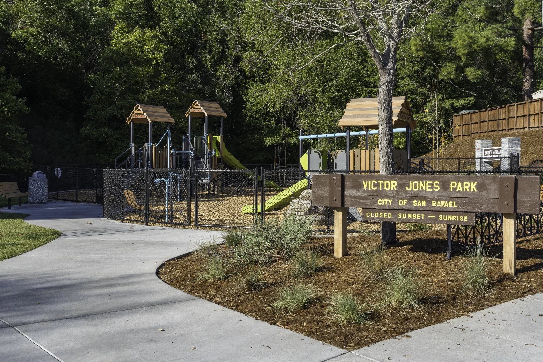 Children's playground with slides and climbing structures, surrounded by trees and a wooden fence, at Victor Jones Park in San Rafael, with a sign indicating park hours from sunset to sunrise.