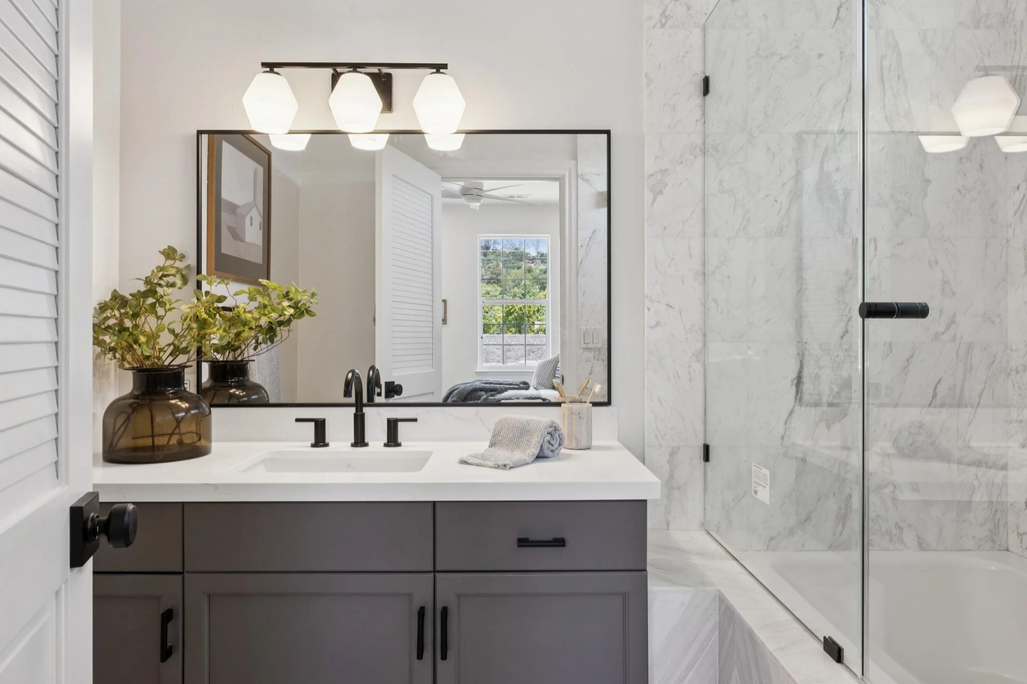 Modern bathroom with a white marble shower, a gray vanity with black hardware, two glass vases with greenery on the sink, and large mirror reflecting a bright bedroom window.