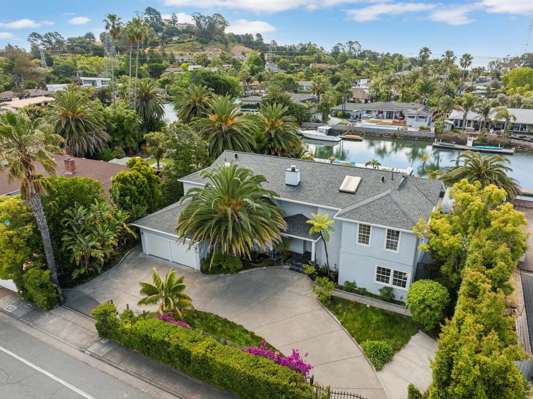 Aerial view of a large white house with a gray roof, surrounded by lush green trees and palm trees, located by a waterway with boats. The neighborhood has other houses, and a hillside with more homes and trees is visible in the background.